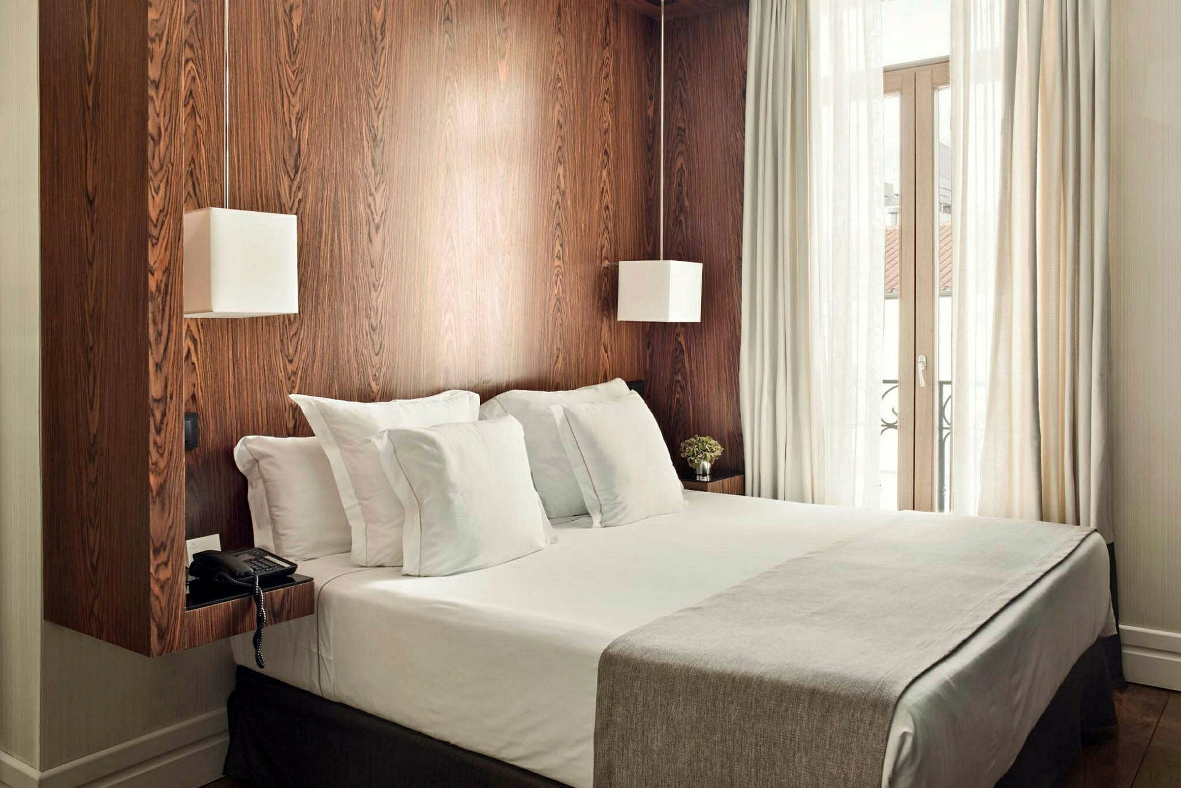 Bild av ett hotellrum med en dubbelsäng, vita sängkläder och träpanel på väggen samt ett stort fönster med ljusa gardiner. (AI-genererad text)