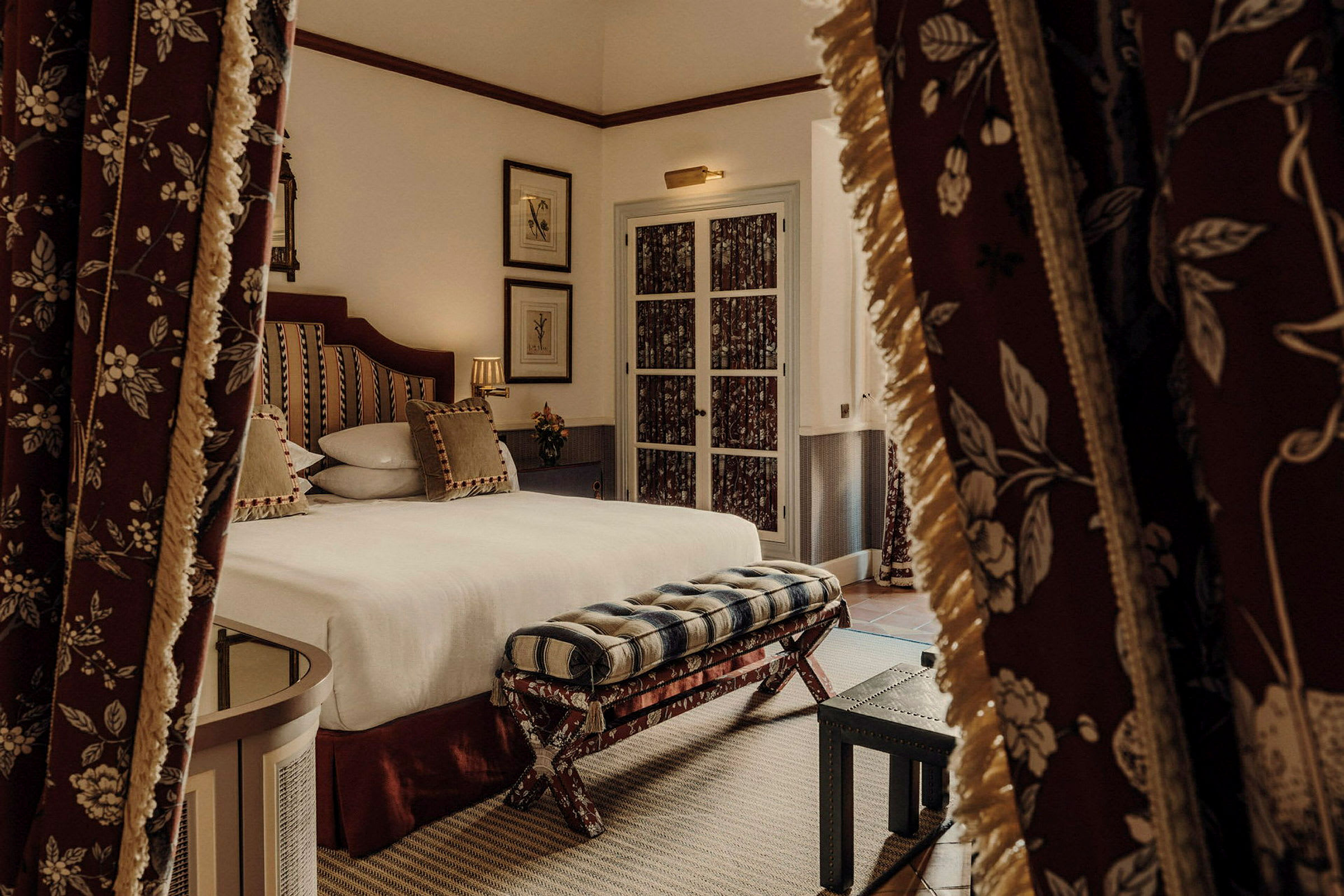 Ett elegant hotellrum med en säng som har dekorativa kuddar och ett fotstöd framför. Rummet har gardiner med blommönster. (AI-genererad text)