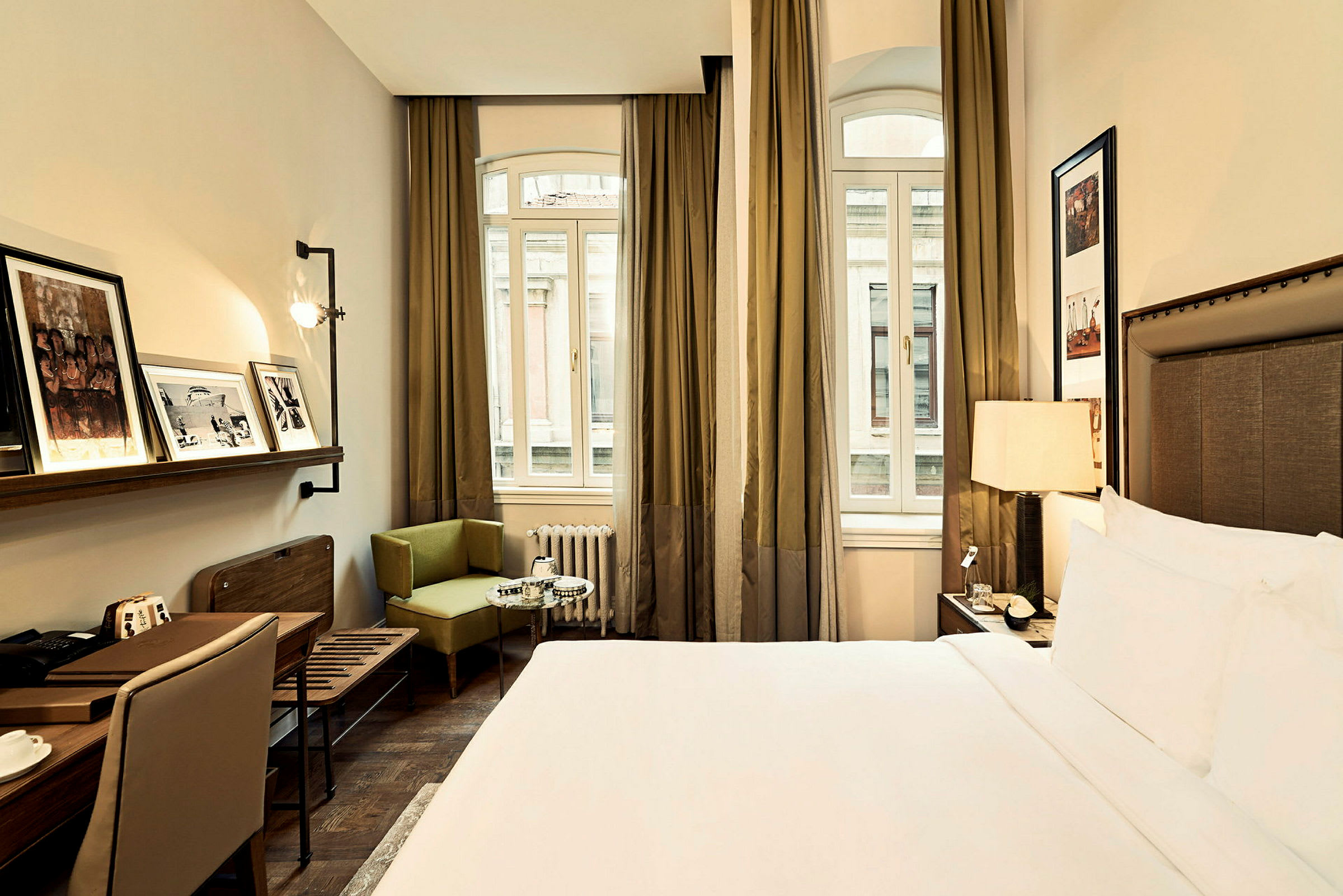 Ett hotellrum med en dubbelsäng, fönster med gardiner och inredning inklusive ett skrivbord, en stol och väggdekorationer. (AI-genererad text)