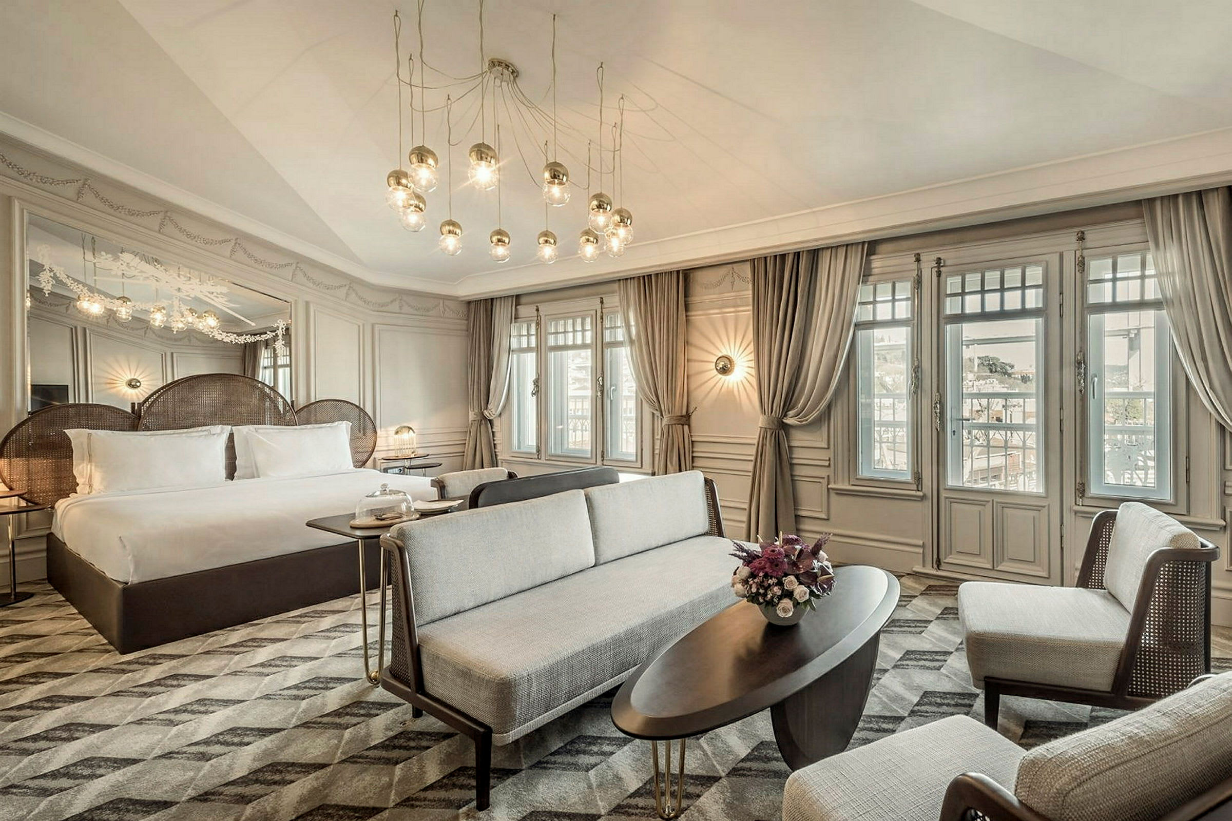 Ett elegant hotellrum med en stor säng, ljuskrona och sittgrupp framför stora fönster. (AI-genererad text)