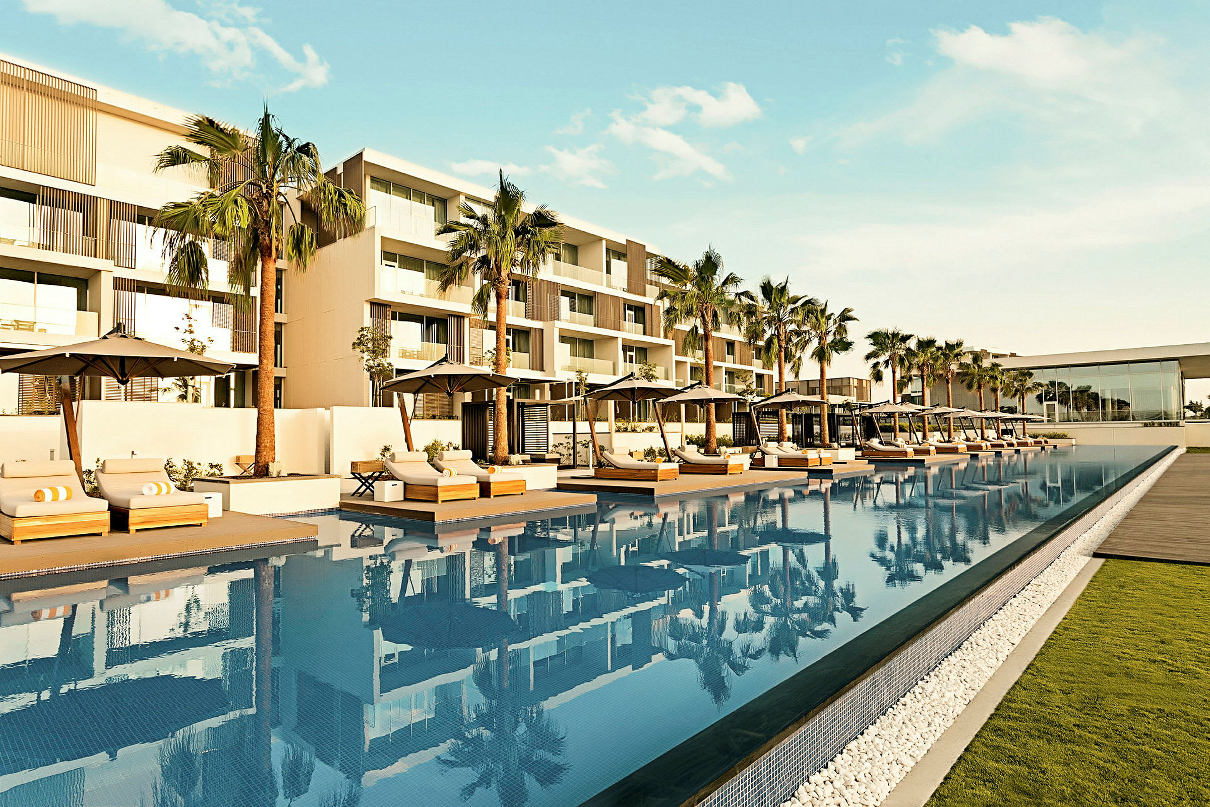 En modern hotellbyggnad med en pool omgiven av solstolar och parasoller i en trädgårdsomgivning. (AI-genererad text)