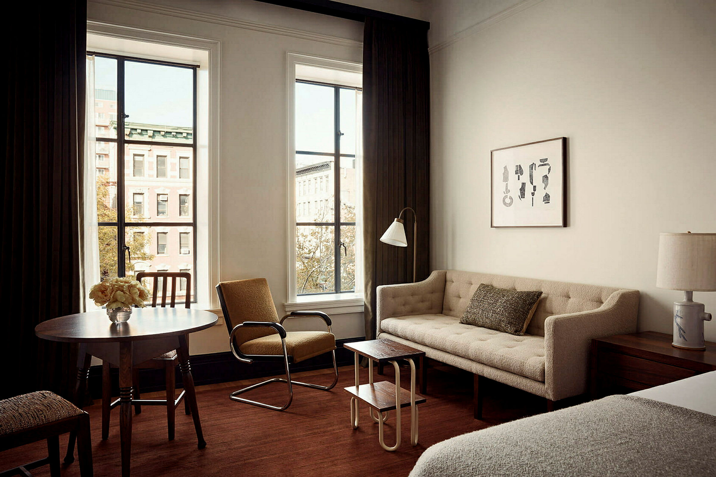 Ett vardagsrum med en beige soffa, en brun fåtölj, ett träbord och två fönster med utsikt mot en byggnad. (AI-genererad text)