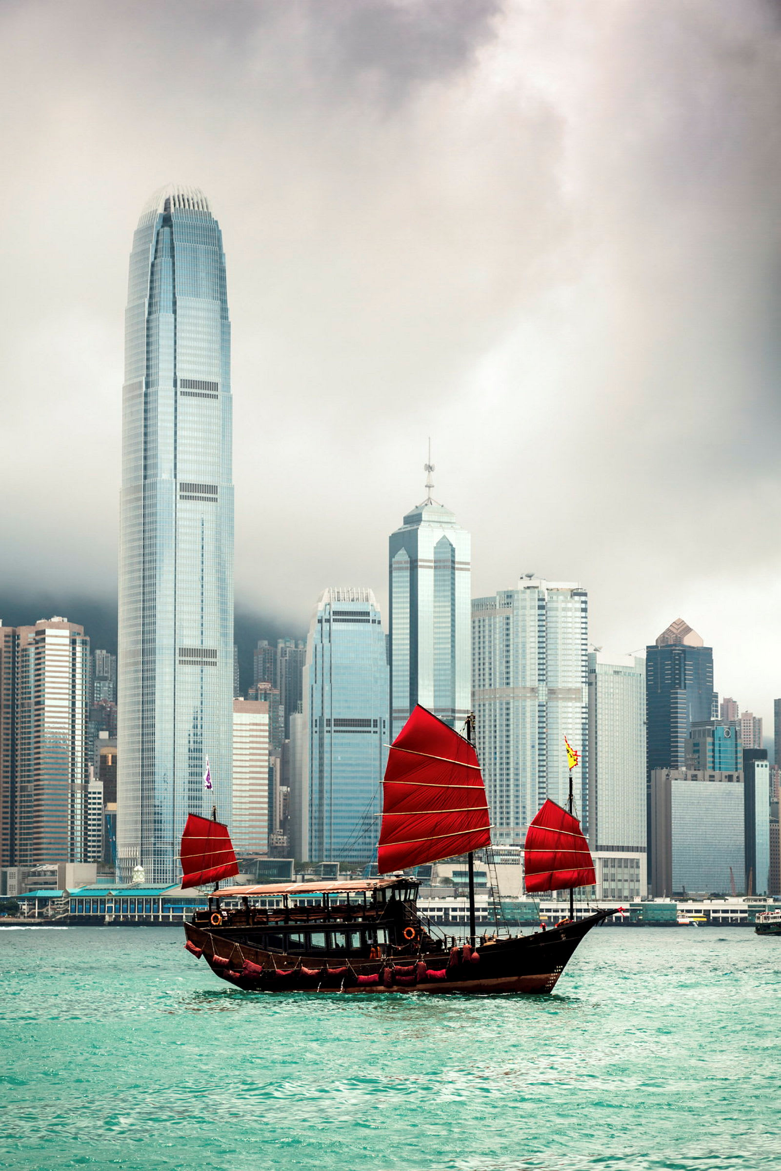 Traditionell kinesisk segelbåt med röda segel framför moderna skyskrapor i Hongkongs hamn. (AI-genererad text)