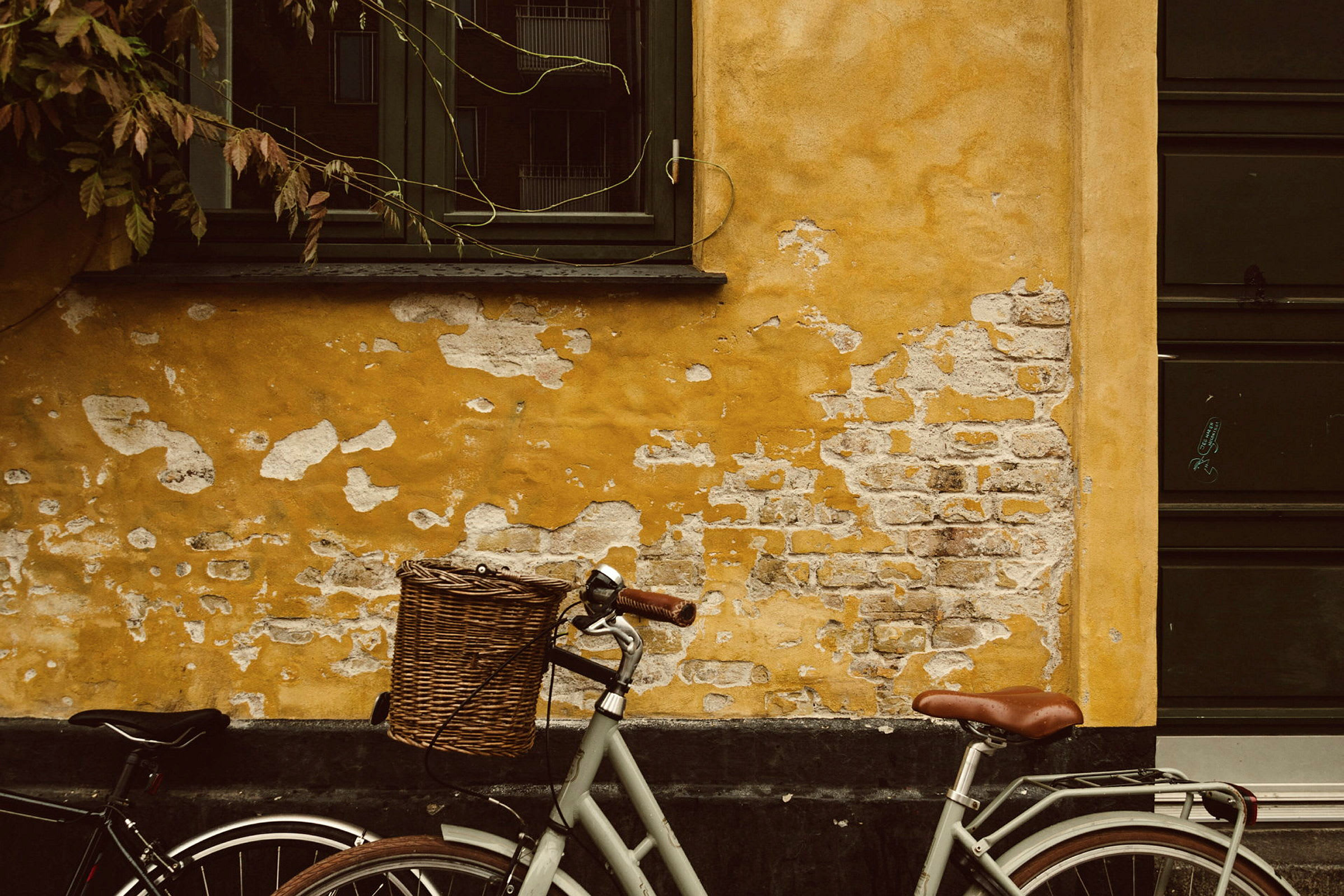 En cykel med brun sadel och korg står lutad mot en gul och slitagen vägg. (AI-genererad text)