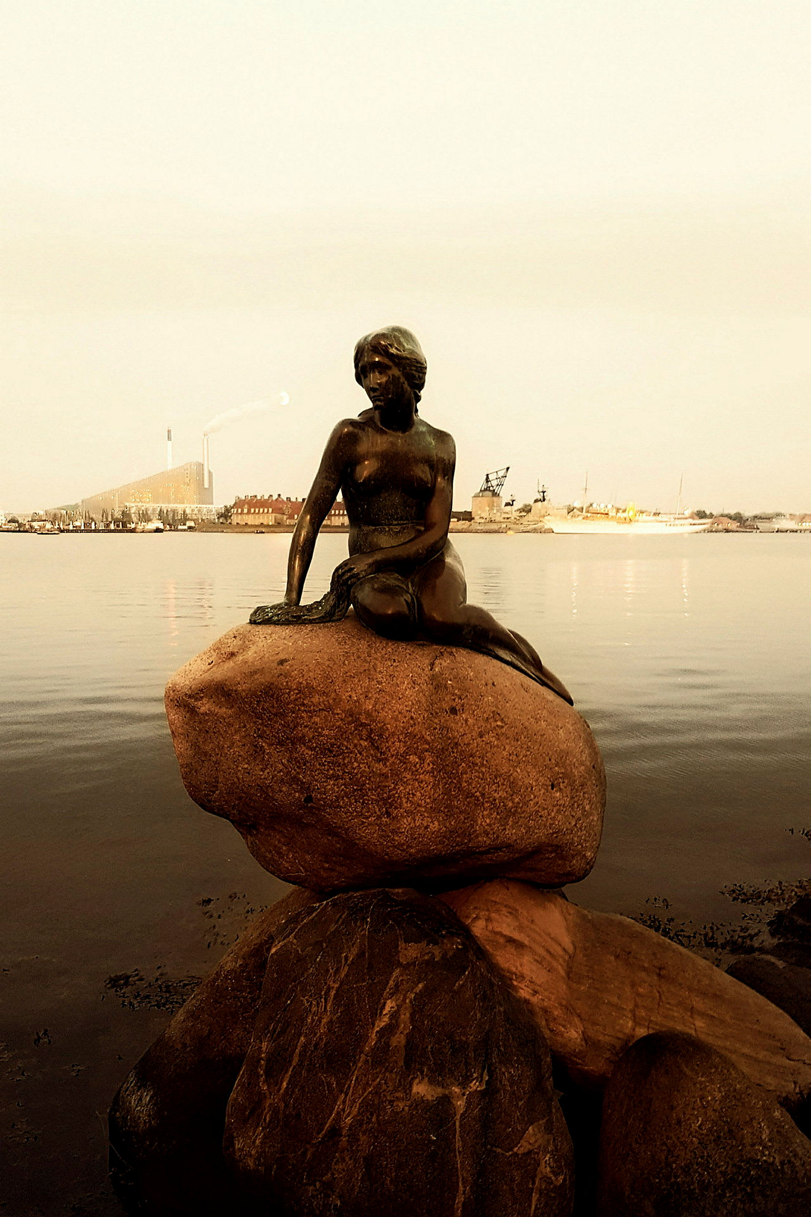 Staty av den lilla sjöjungfrun på en sten vid vattnet med en stadsbild i bakgrunden. (AI-genererad text)