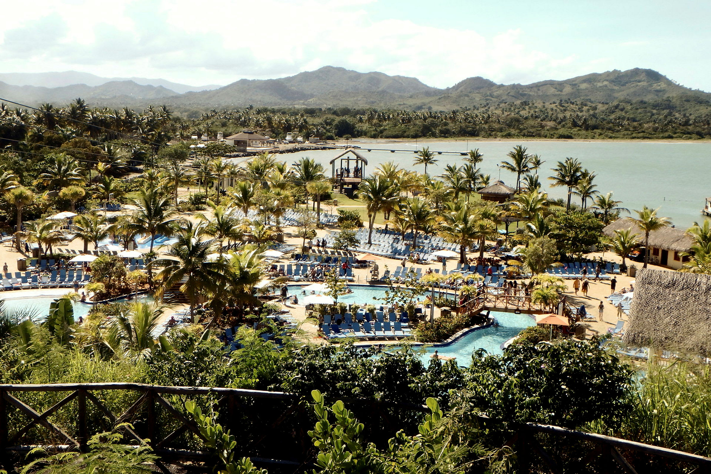 En utsikt över en resort med flera pooler, palmer och solstolar nära ett vattenlandskap. (AI-genererad text)