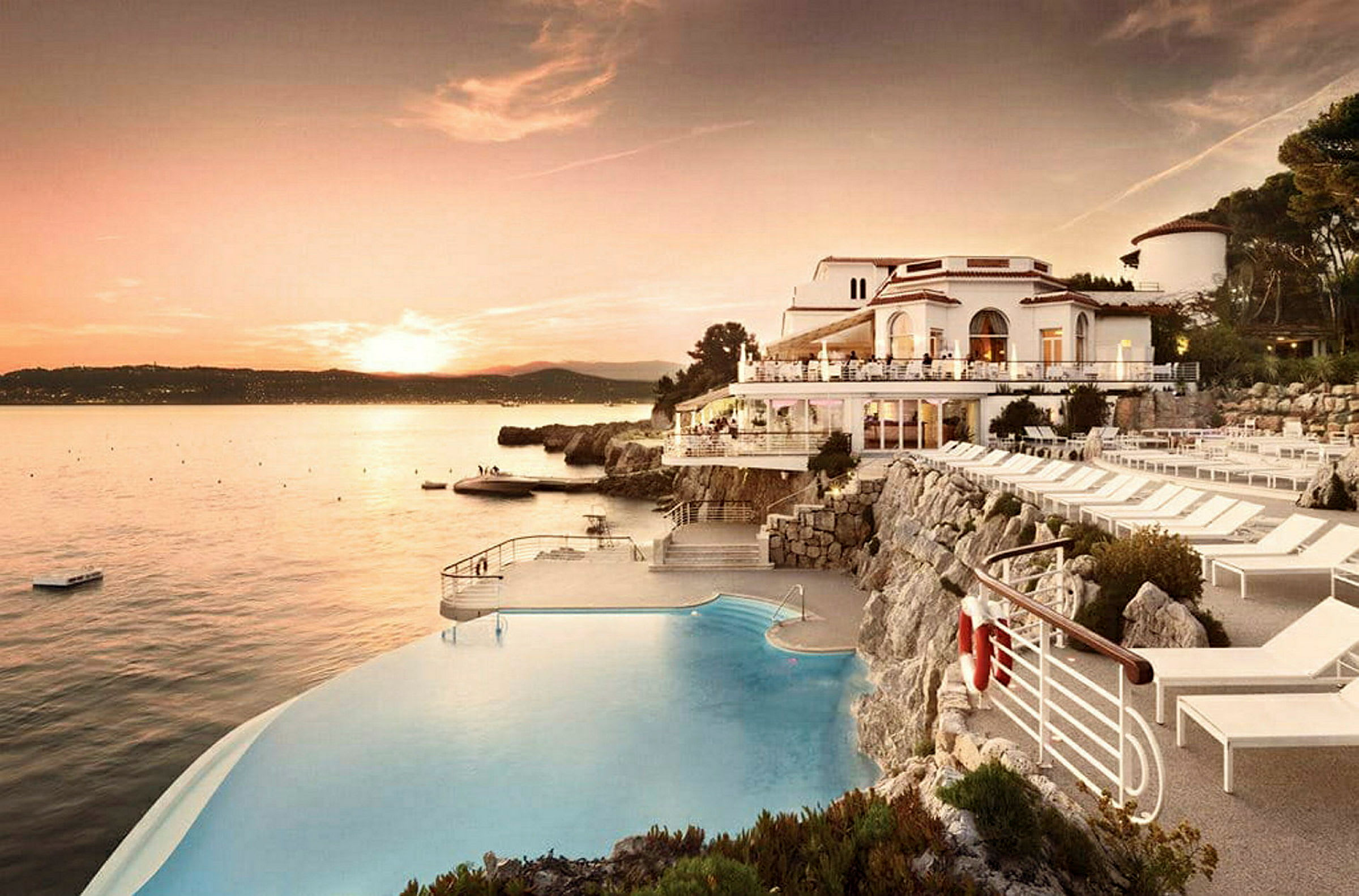 Vy över havet vid solnedgång med en infinitypool och vita byggnader vid klippkanten. (AI-genererad text)