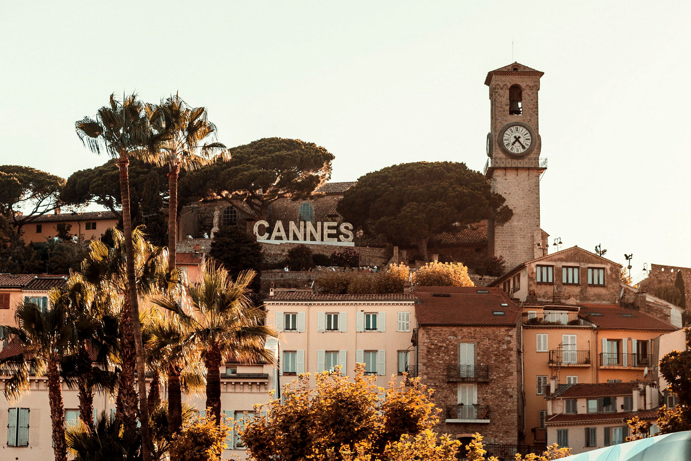 Utsikt över Cannes med en historisk klocktorn och skylt med stadens namn, omgiven av träd och byggnader. (AI-genererad text)
