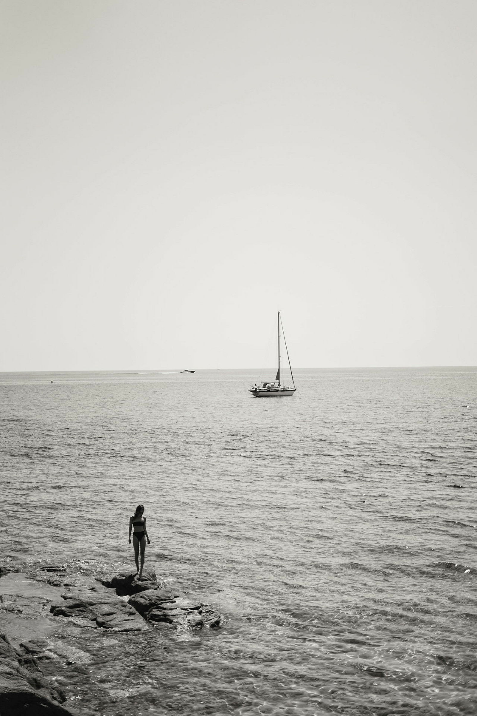 En person står på en klippa nära havet med en segelbåt i bakgrunden. (AI-genererad text)