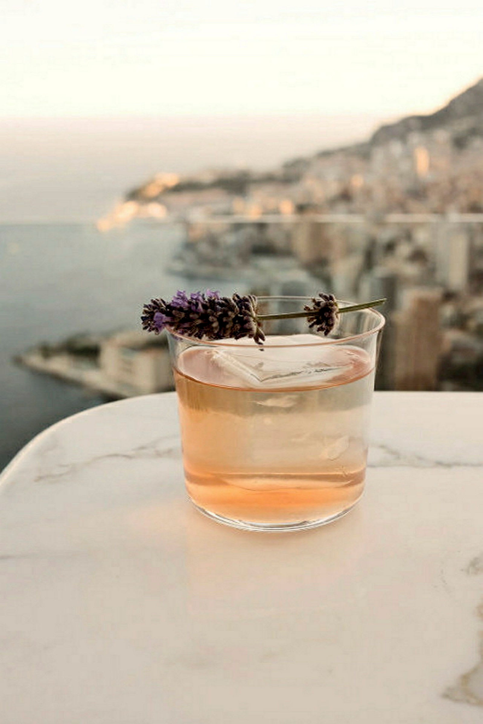 Ett glas med en ljus vätska garnerat med lavendel, placerat på ett marmorbord med en utsikt över en kuststad. (AI-genererad text)