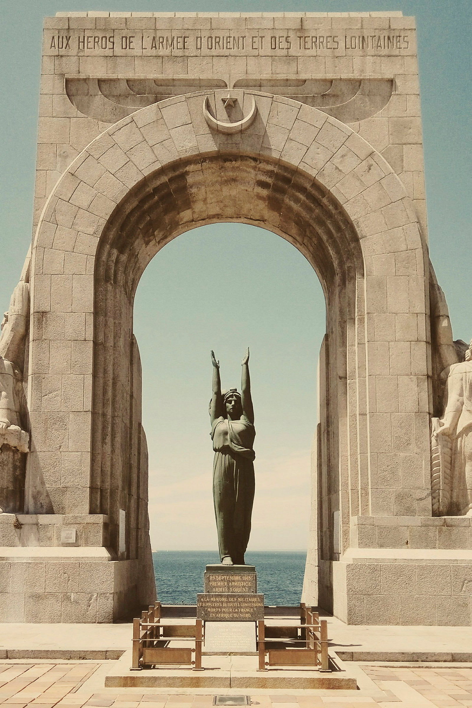 Ett monument i sten med en båge och en staty av en kvinna med armarna upplyfta, placerad vid havet. (AI-genererad text)