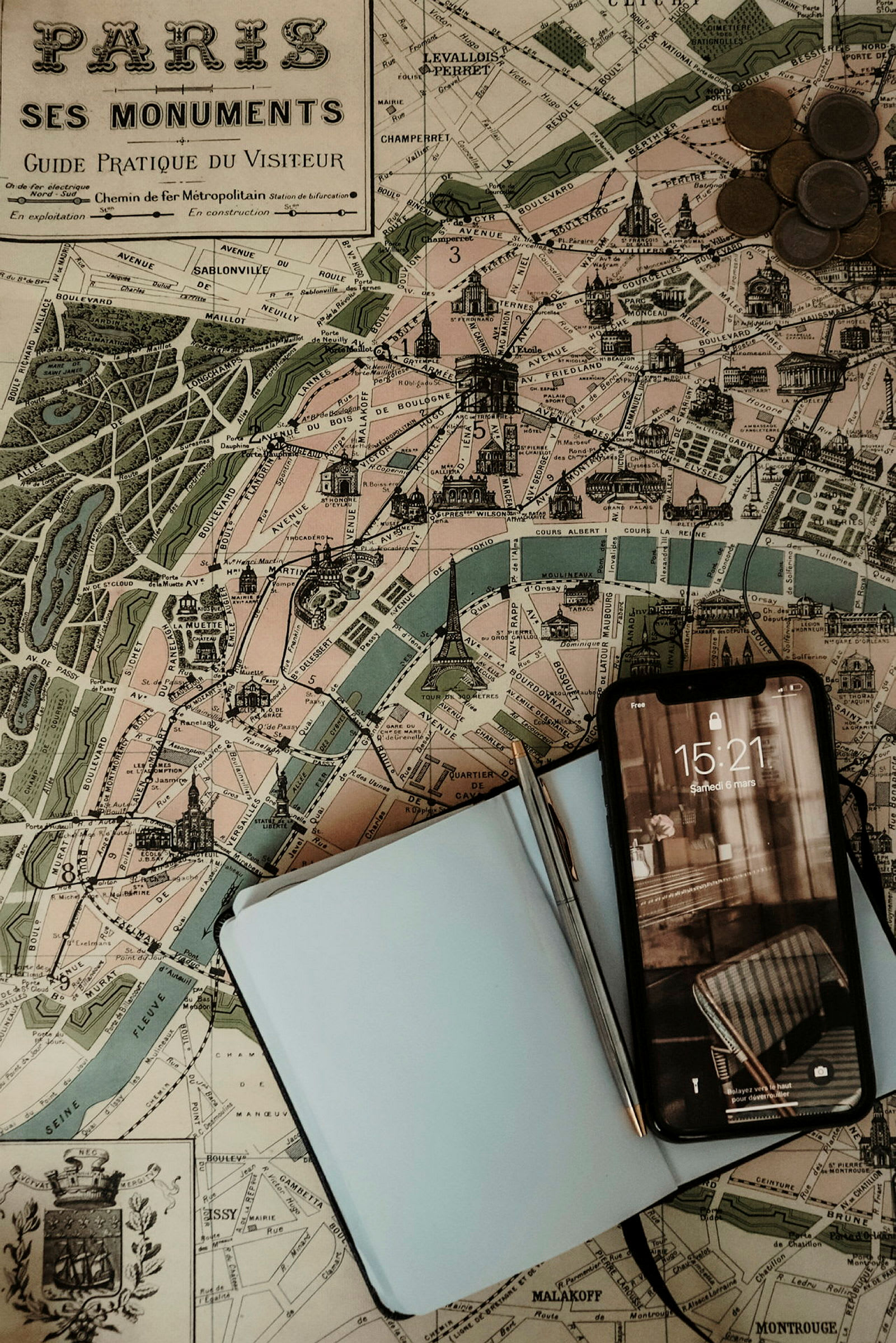 En bild av en vintage karta över Paris. På kartan ligger en anteckningsbok, en penna och en mobiltelefon samt några mynt. (AI-genererad text)