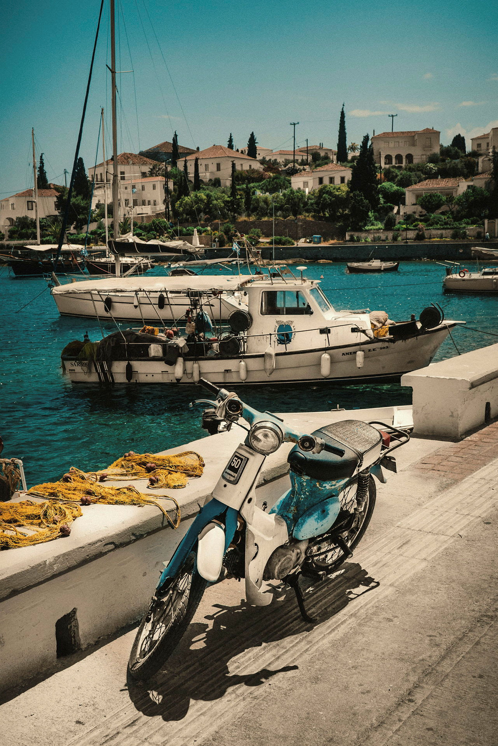 En blå och vit moped står parkerad vid en kaj, med fiskebåtar och vattnet i bakgrunden. (AI-genererad text)