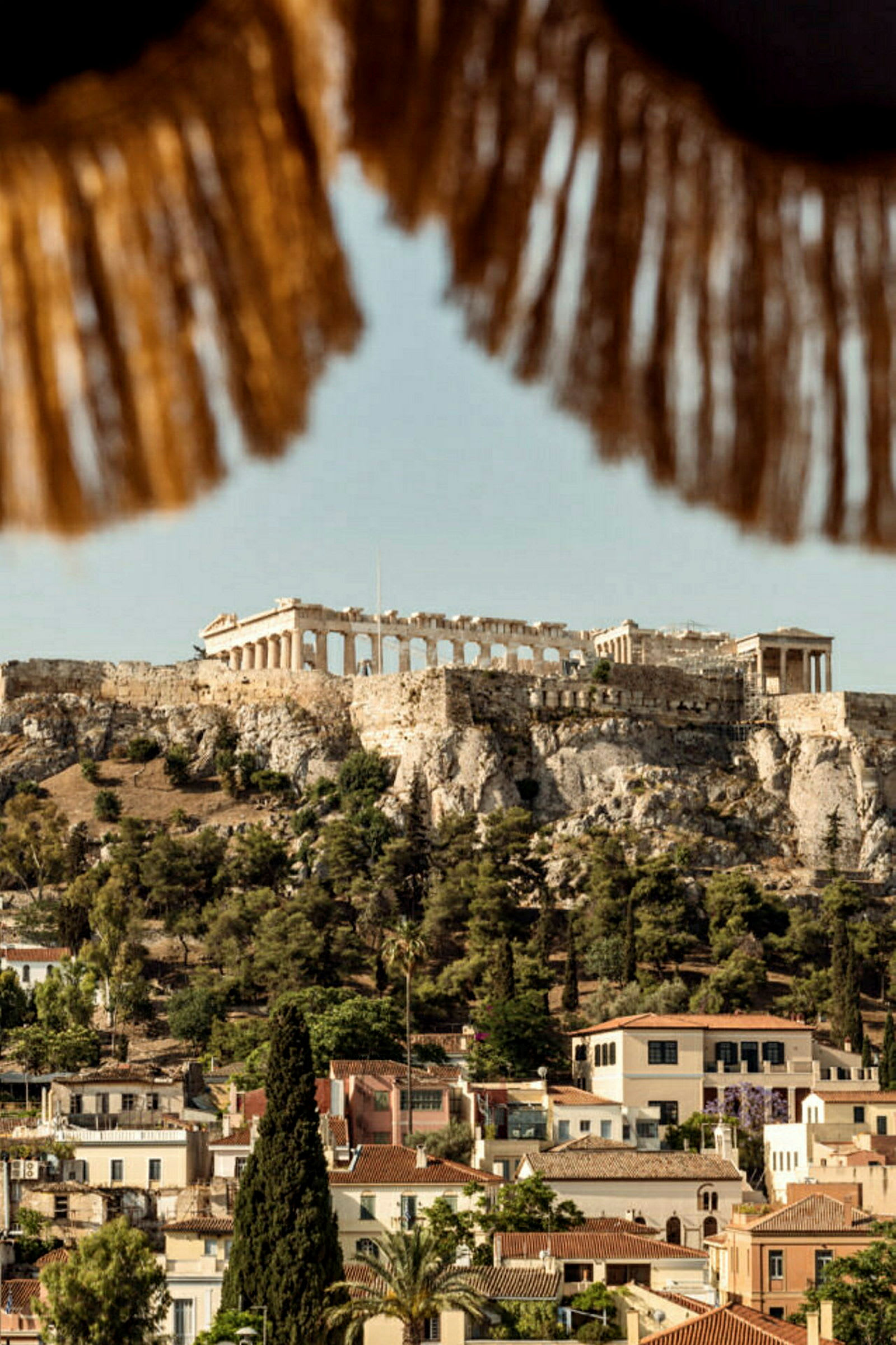Vy av Akropolis i Aten med omgivande byggnader och vegetation, med delar av ett parasoll synligt i förgrunden. (AI-genererad text)