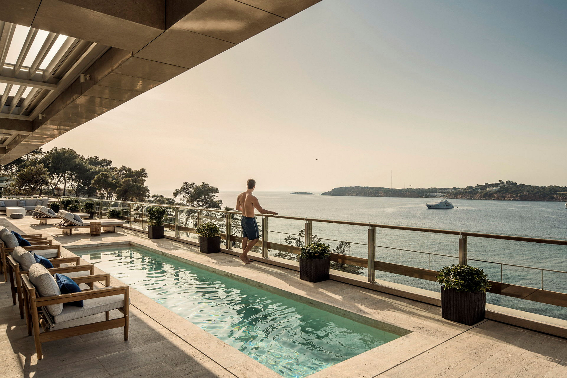 En person står vid balkongräcket med utsikt över havet. Mjukstolar och en pool är placerade på terrassen. (AI-genererad text)
