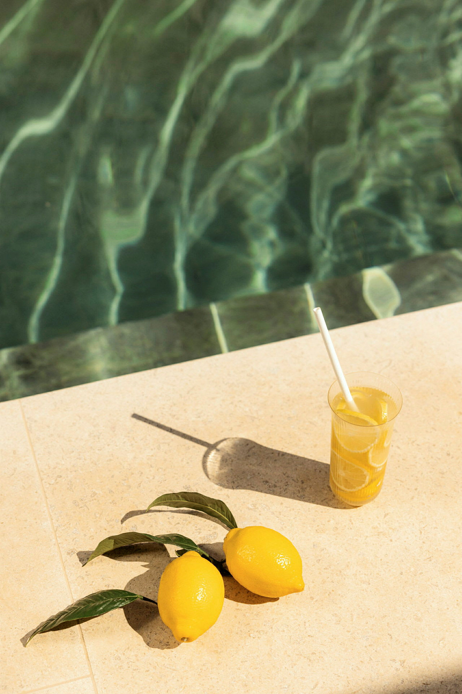 Ett glas med citrondryck och två citroner placerade bredvid en pool. (AI-genererad text)