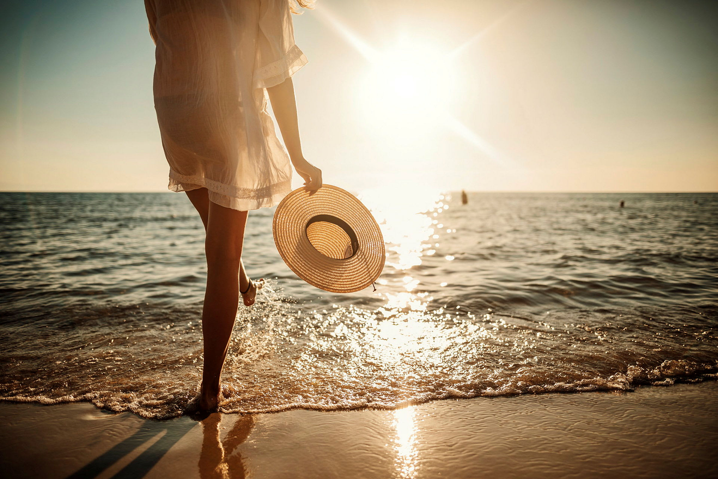 En person går vid strandkanten med en solhatt i handen vid solnedgången. (AI-genererad text)