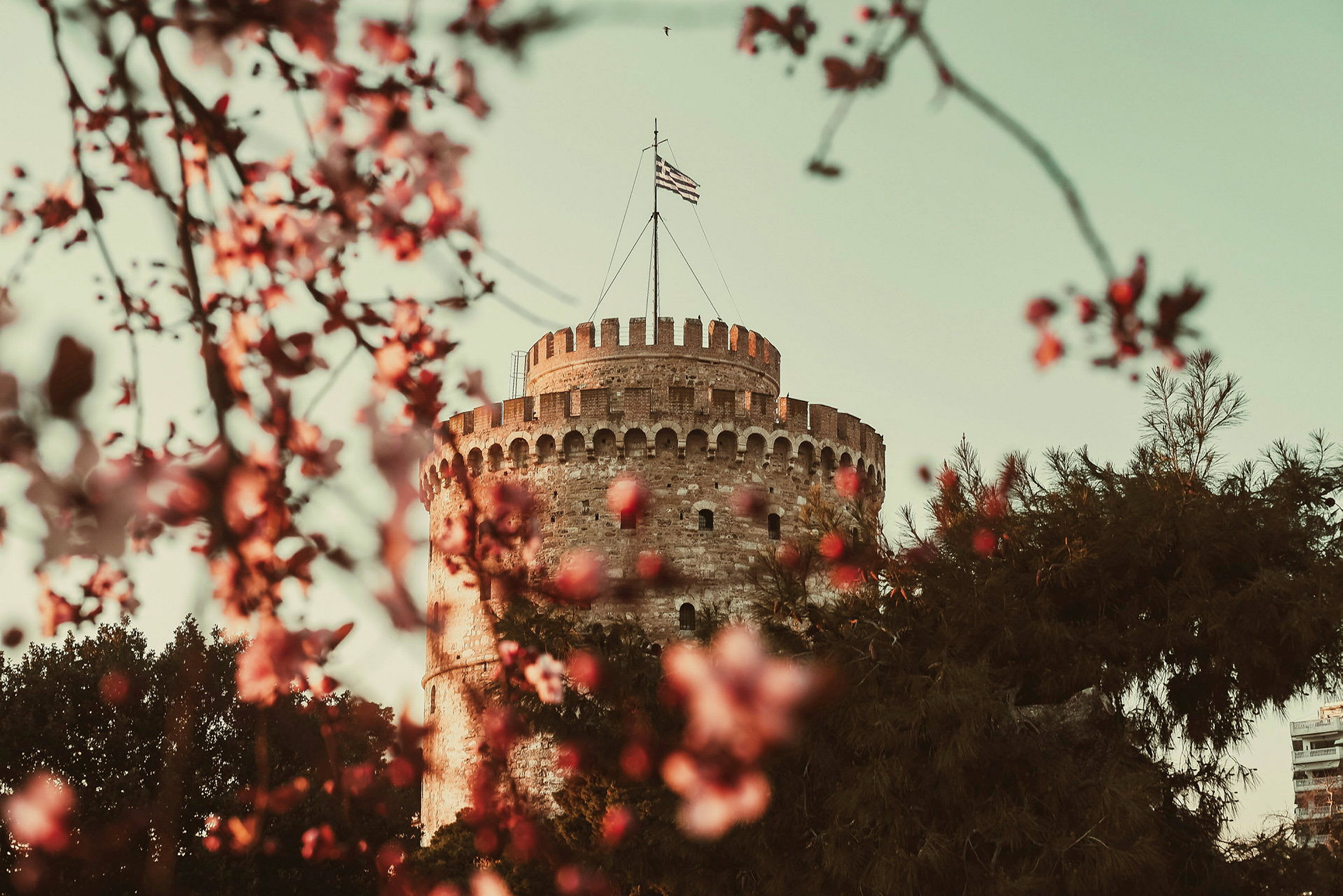 Vit torn byggnad med en grekisk flagga på toppen omgiven av blommor och träd. (AI-genererad text)