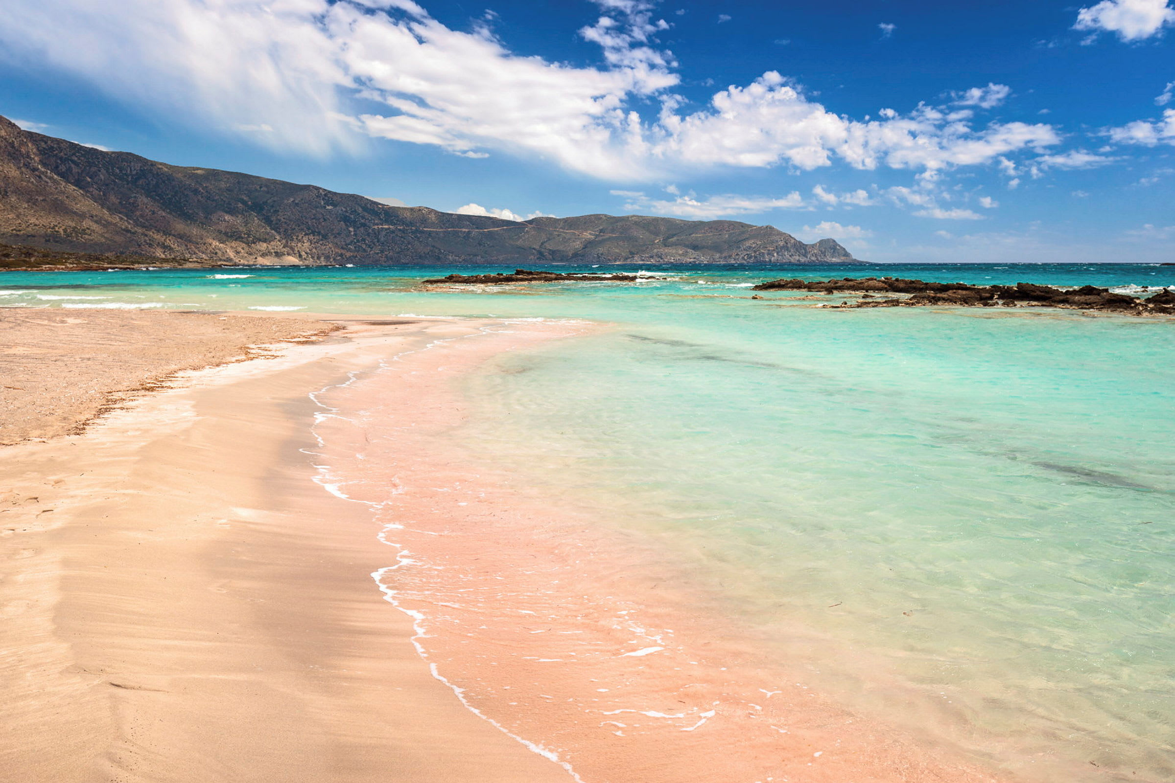 En strand med rosa sand och klart turkost vatten, omgiven av klippor och berg i fjärran. (AI-genererad text)