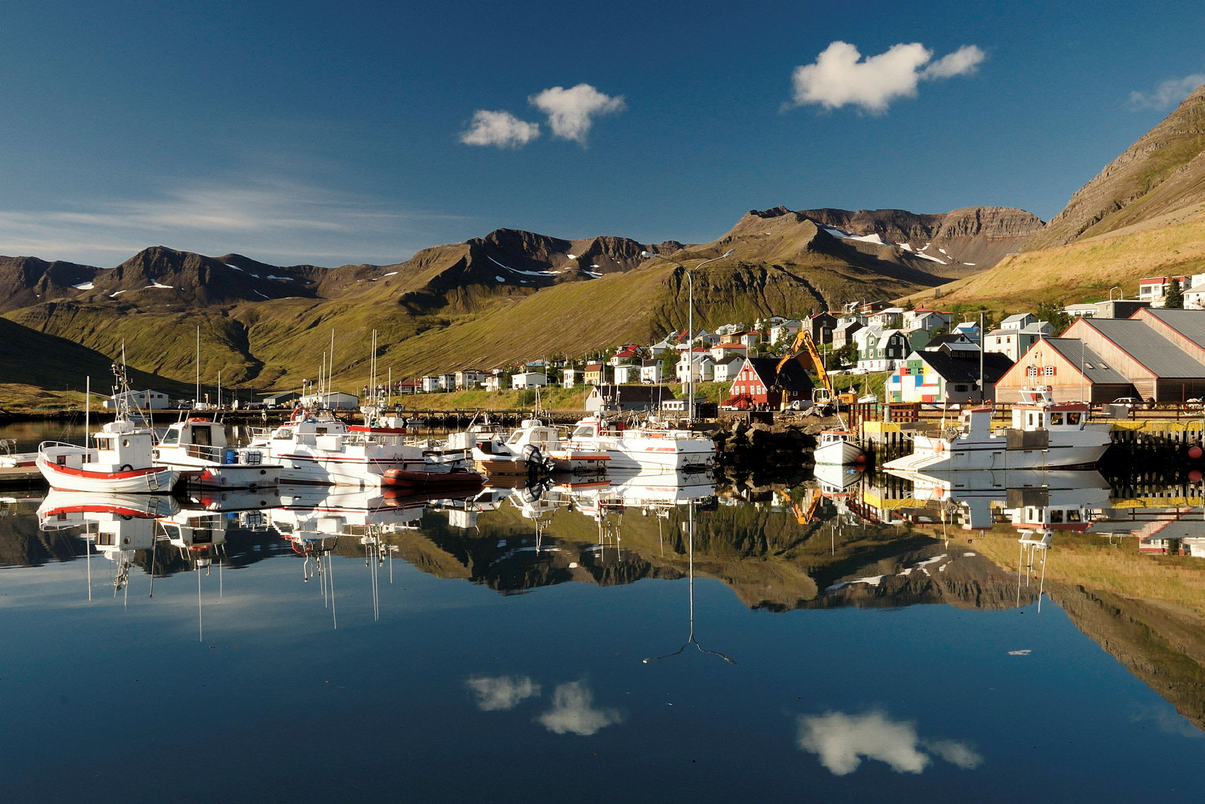 Båtar vid en hamn med byggnader och berg i bakgrunden. (AI-genererad text)