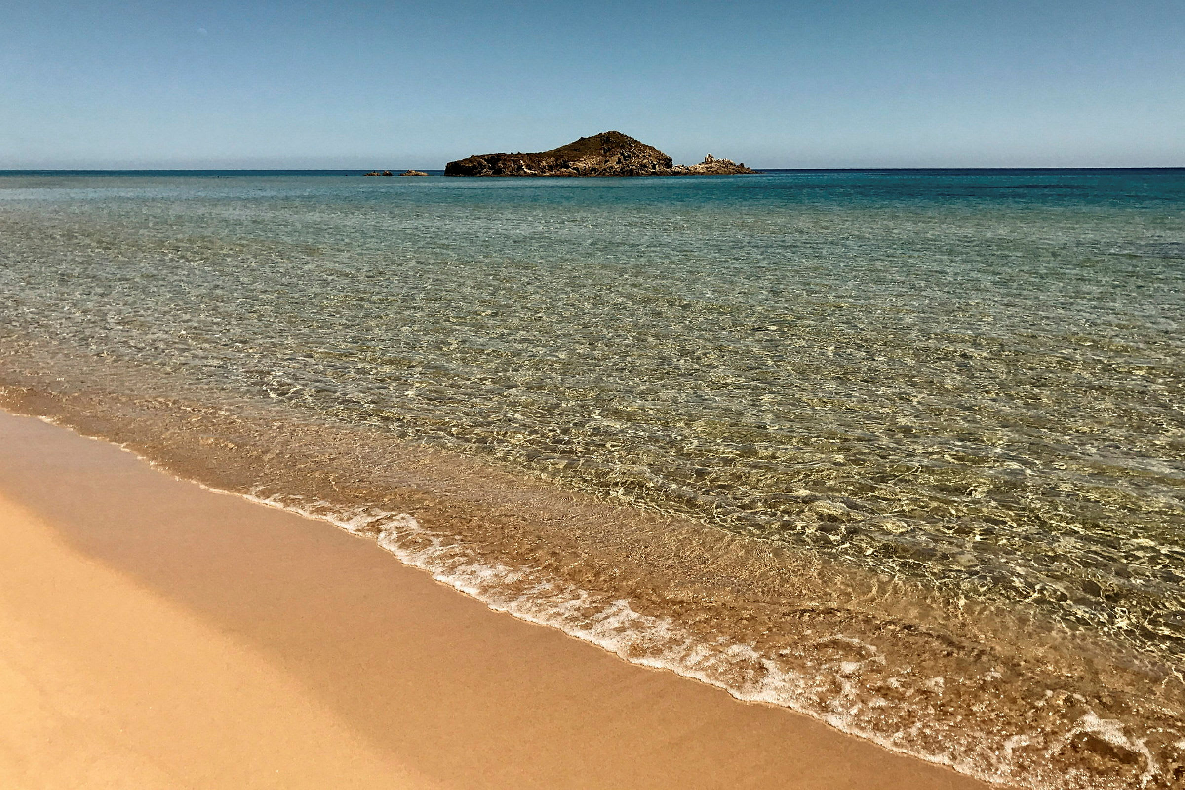 Strand med klarblått vatten och en liten ö i bakgrunden. (AI-genererad text)