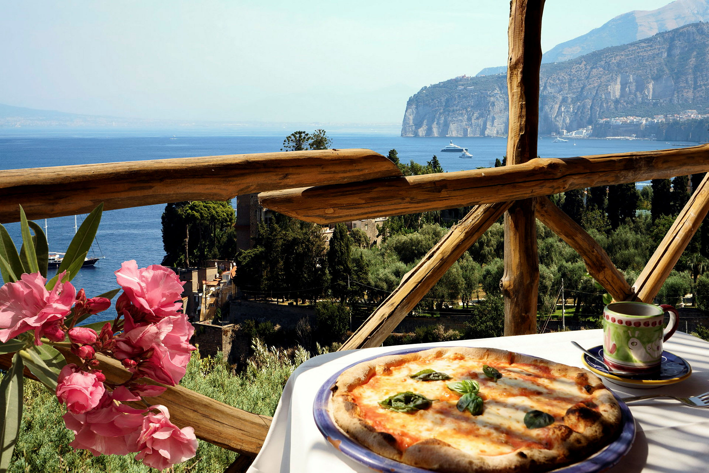 En pizza på ett bord med havsutsikt, träräcke och rosa blommor i förgrunden. (AI-genererad text)