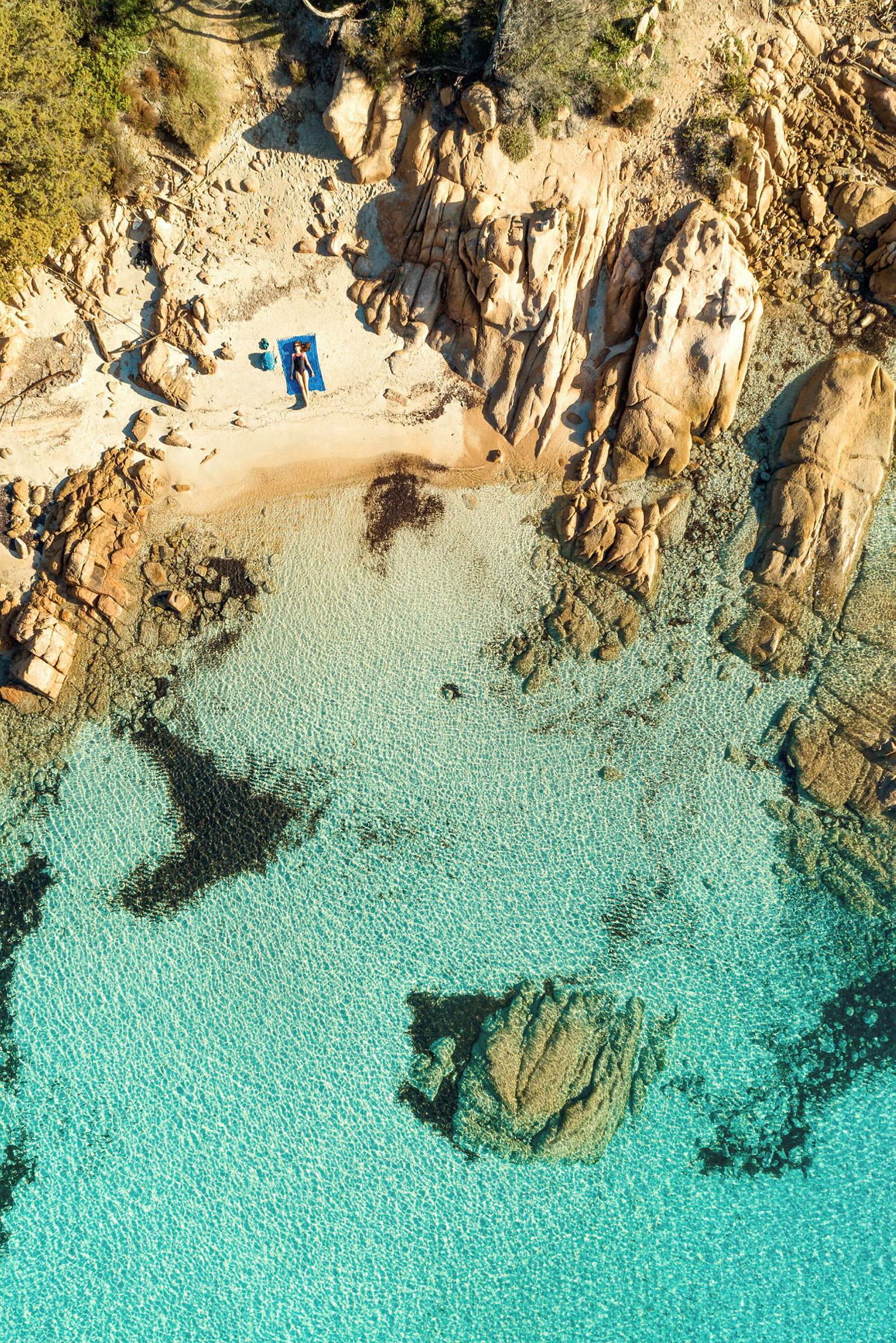 Flygfoto av en liten strand med en person på en blå handduk nära klippor, omgiven av klart turkost vatten. (AI-genererad text)