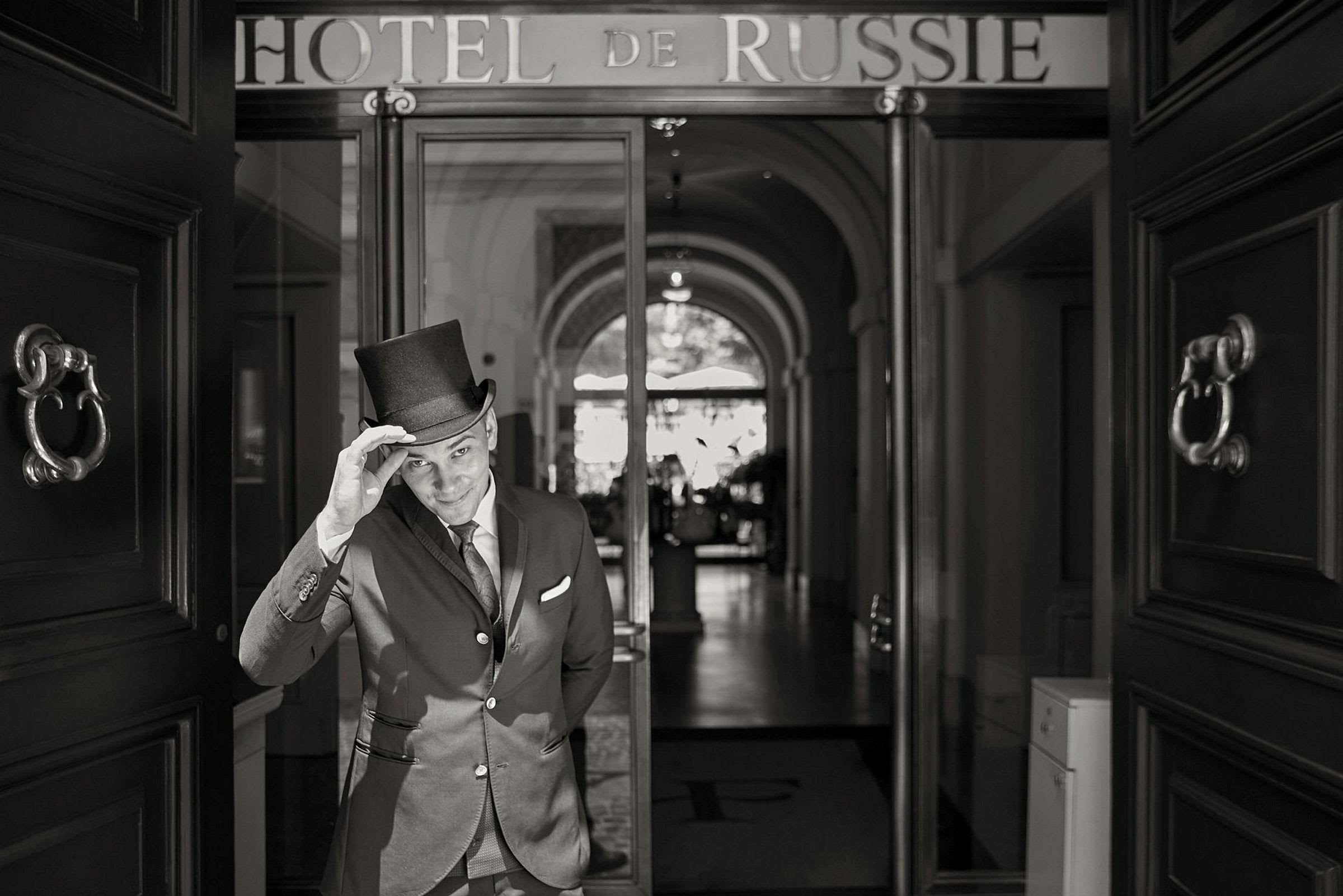 En person i kostym med cylinderhatt står vid ingången till ett hotell med texten 'Hotel de Russie' ovan. (AI-genererad text)