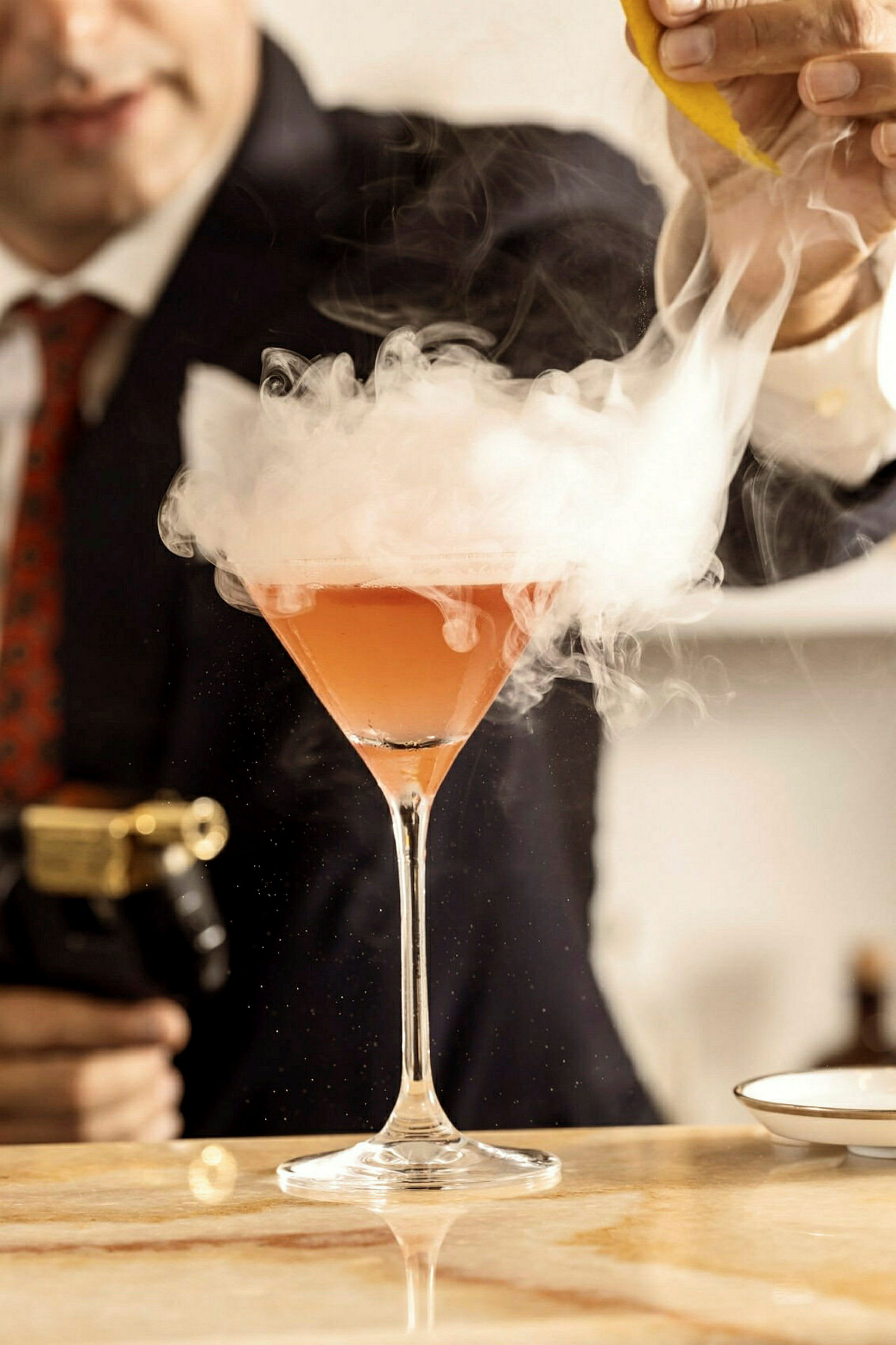 En cocktail i ett martiniglas med rök ovanför, en person håller en citronskal ovanför glaset. (AI-genererad text)