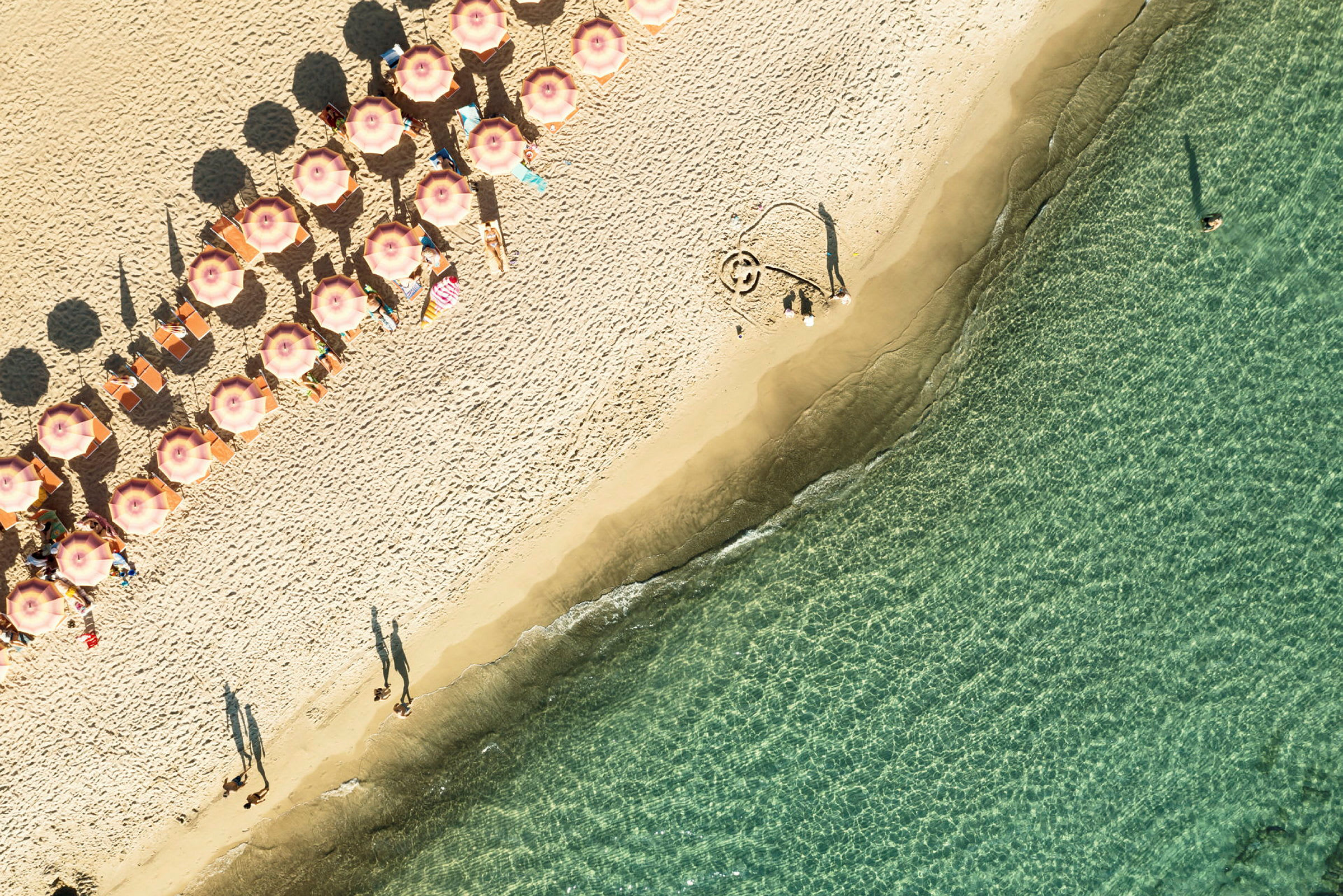 Luftvy över en strand med solstolar, parasoller och personer vid vattnet. (AI-genererad text)