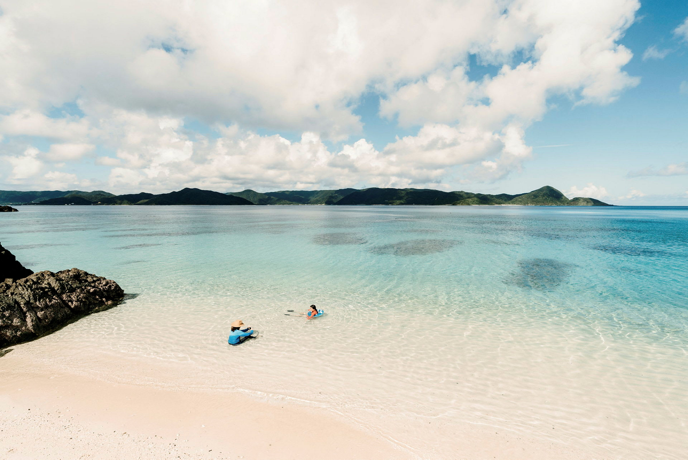 En strand med vit sand vid ett klart blått hav, med två personer som sitter och simmar nära vattenkanten. (AI-genererad text)
