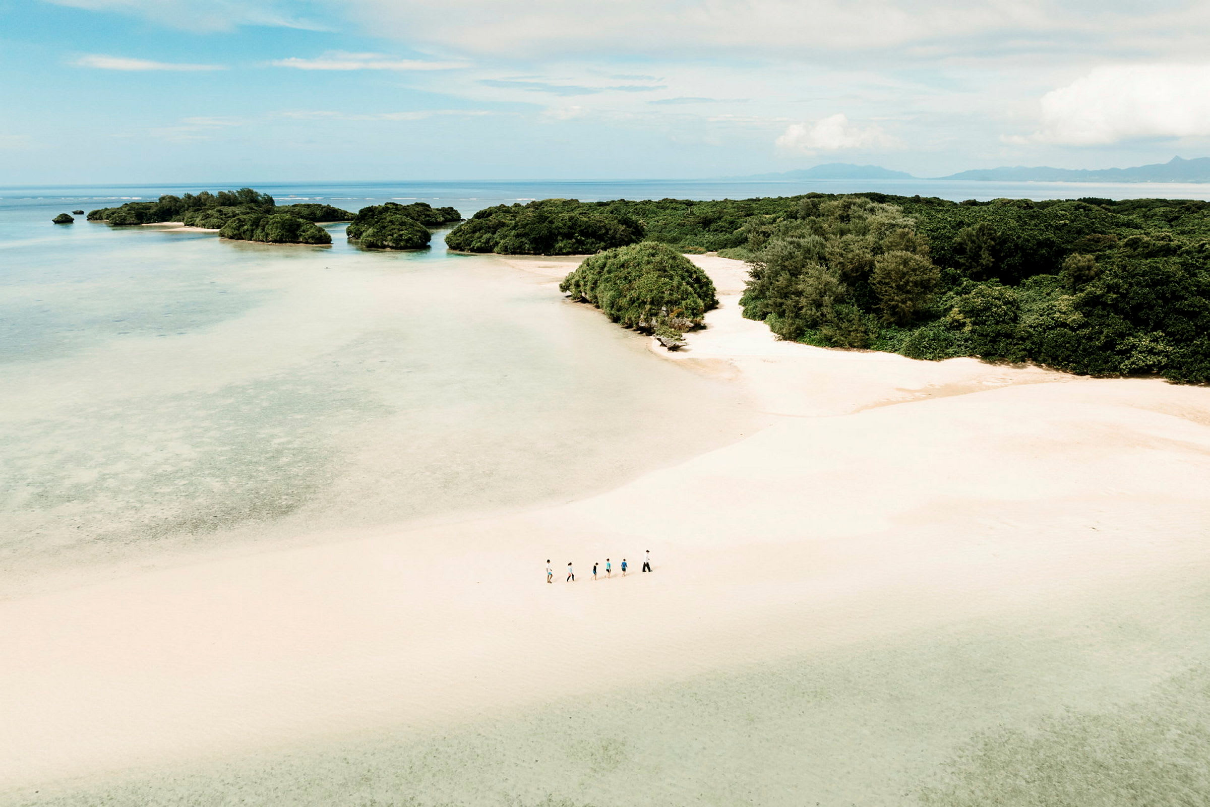 En flygbild av en sandstrand med gröna buskar och omgivande små öar i ett klart, blått hav. En grupp människor går längs stranden. (AI-genererad text)