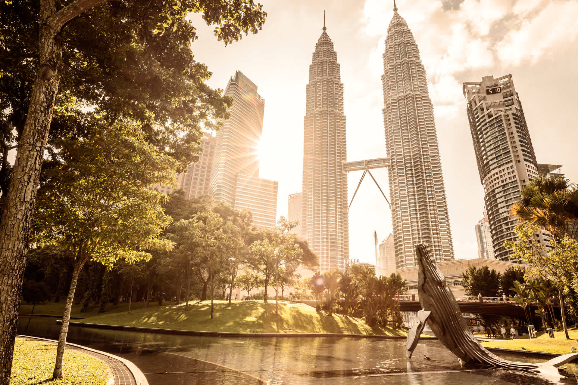 Utsikt över Petronas Twin Towers med solnedgång, gröna träd och en skulptur föreställande en val i förgrunden. (AI-genererad text)