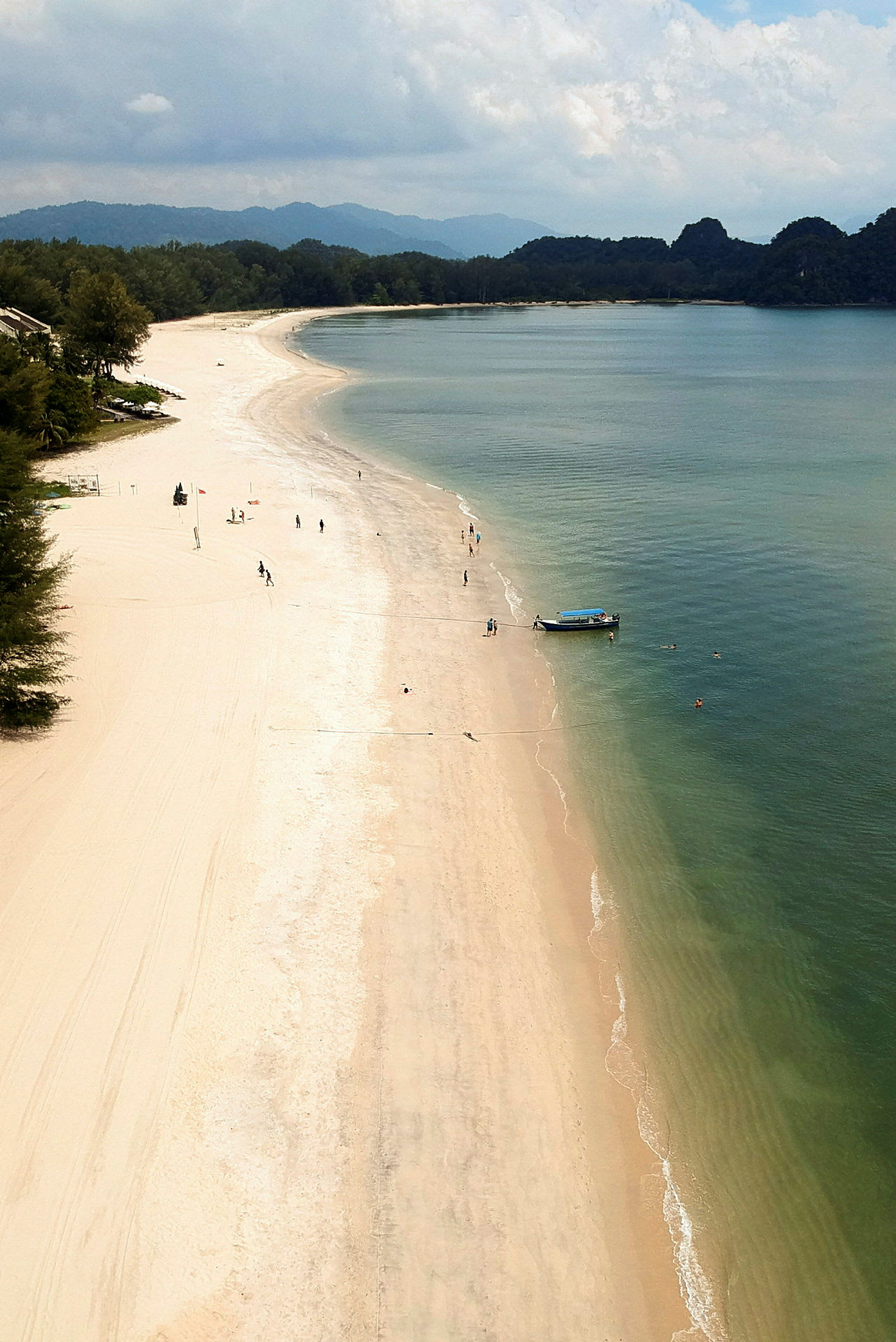 Vy över en lång sandstrand med klart vatten och några människor och en båt nära vågornas kant. (AI-genererad text)