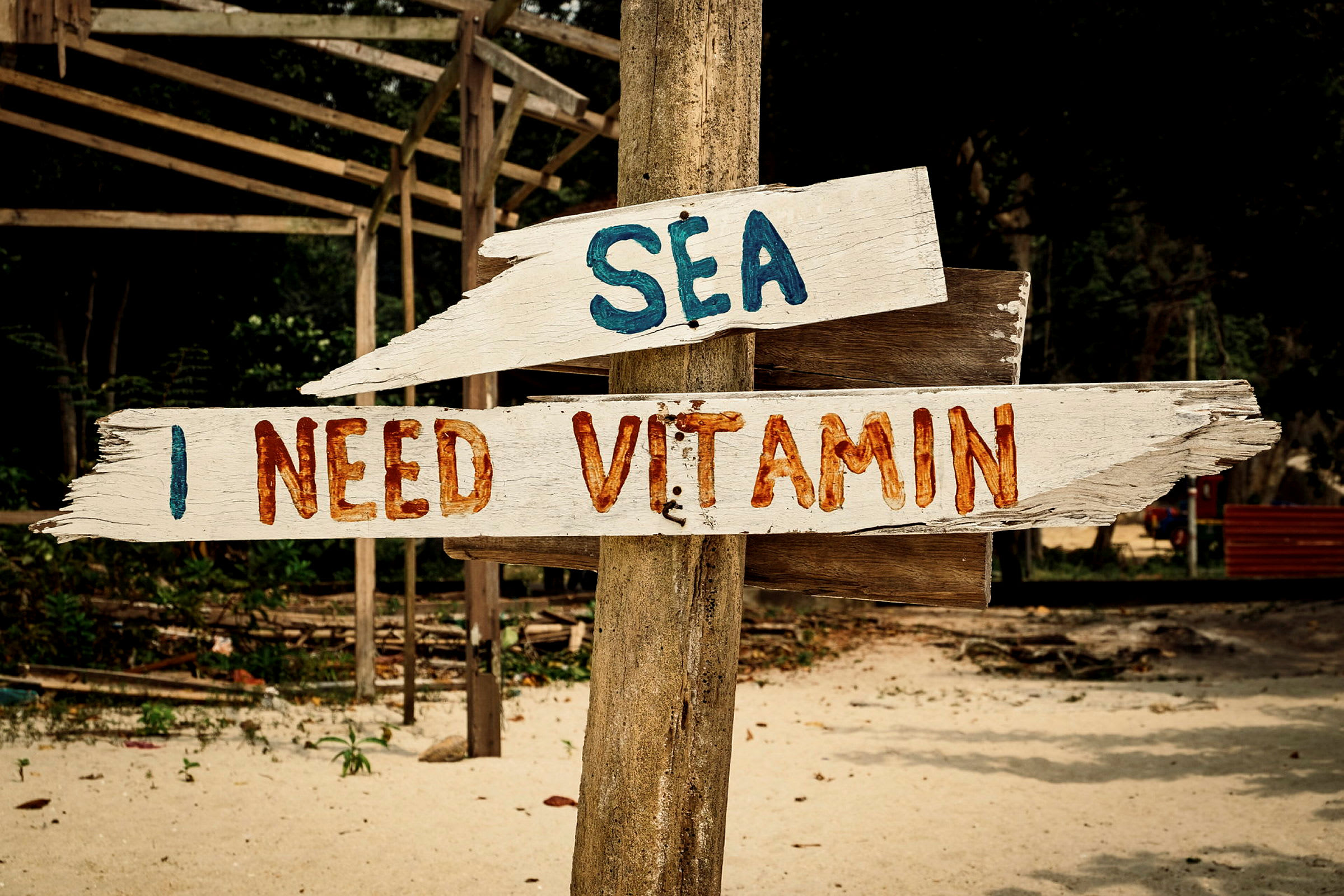Träskyltar med texten 'SEA' och 'I NEED VITAMIN' på en strand med bakgrund av träd. (AI-genererad text)