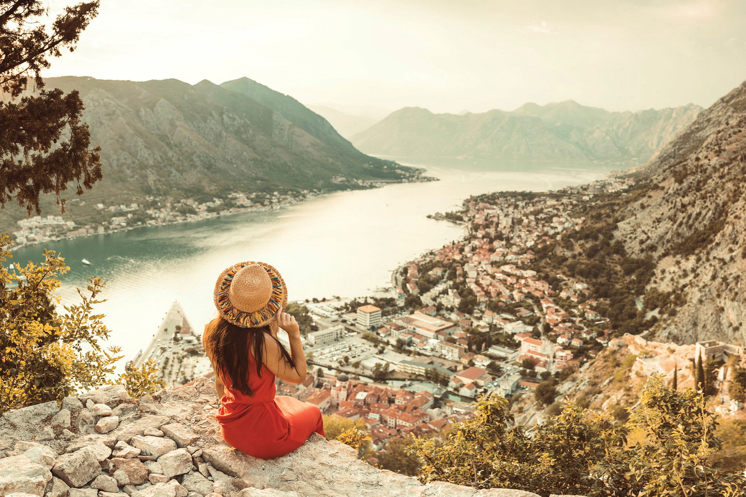Person sittande på en klippa med utsikt över en stad vid vattnet och omgivande berg. (AI-genererad text)