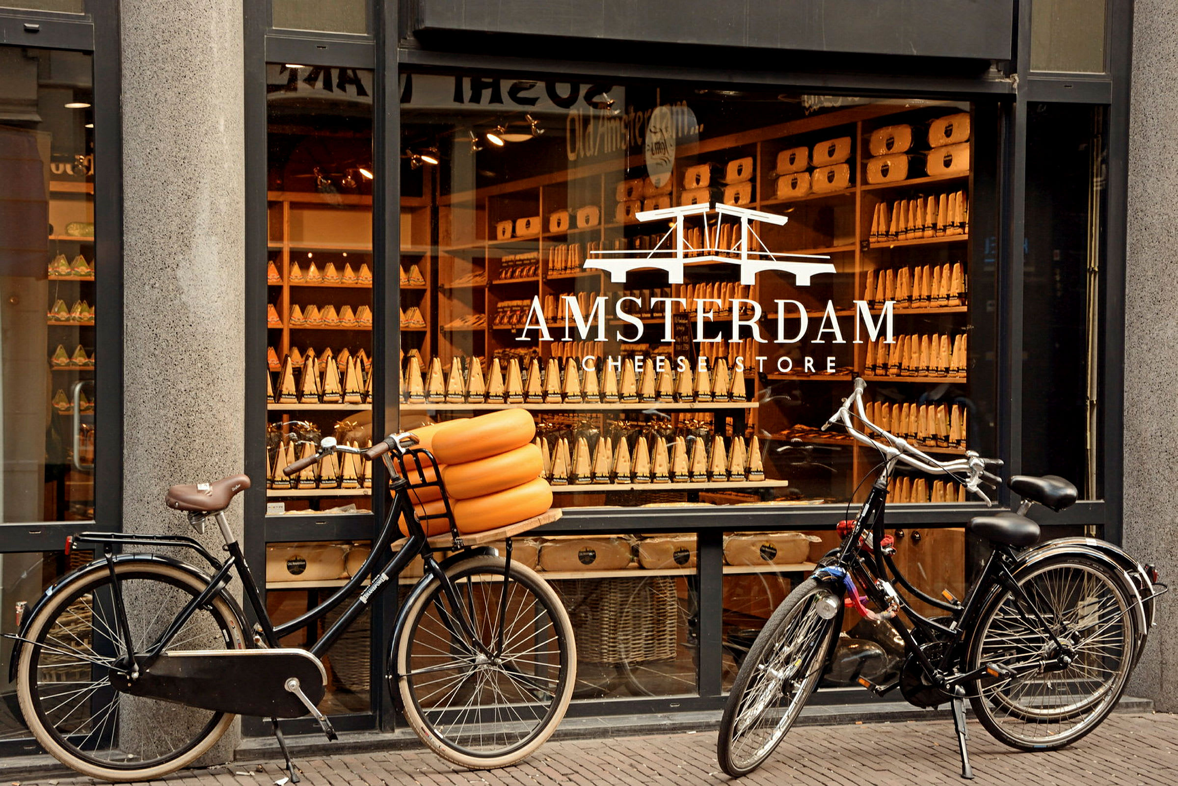Två cyklar framför en butik med texten 'Amsterdam Cheese Store' på fönstret. (AI-genererad text)