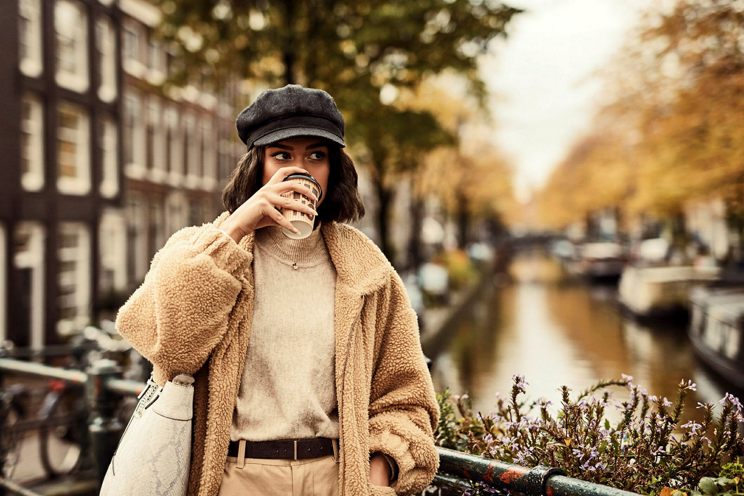 En person klädd i beige står vid en kanal med blommor och byggnader i bakgrunden. (AI-genererad text)