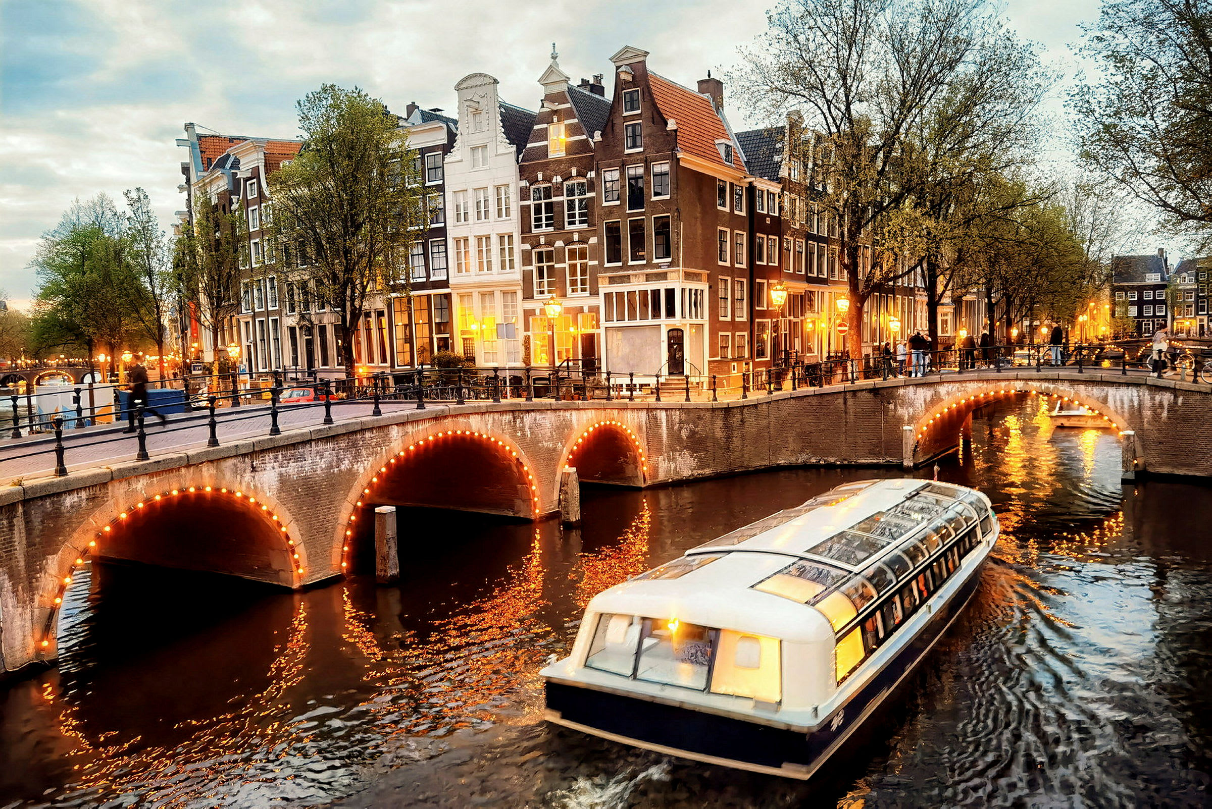 Vy över en kanal i Amsterdam med en båt i förgrunden och upplysta broar samt byggnader. (AI-genererad text)