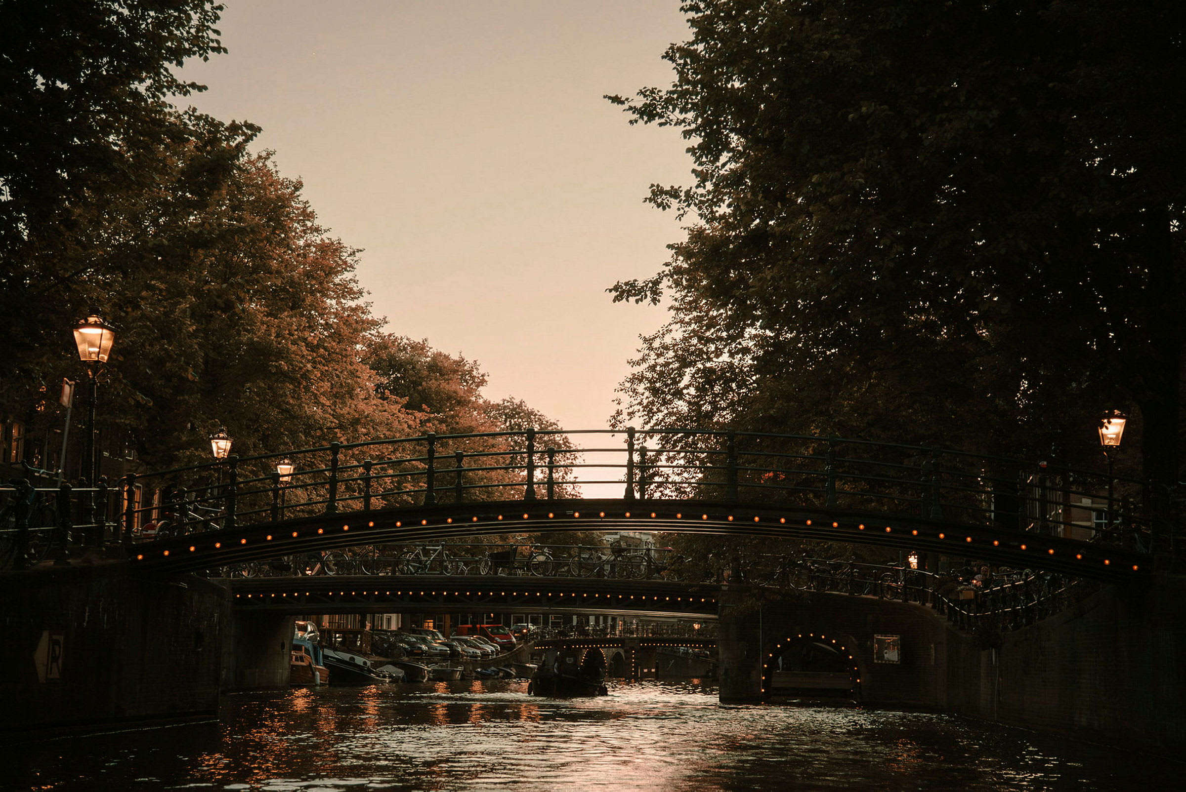 Bro över en kanal i stadsmiljö under skymning, omgiven av lövträd och gaturlampor. (AI-genererad text)