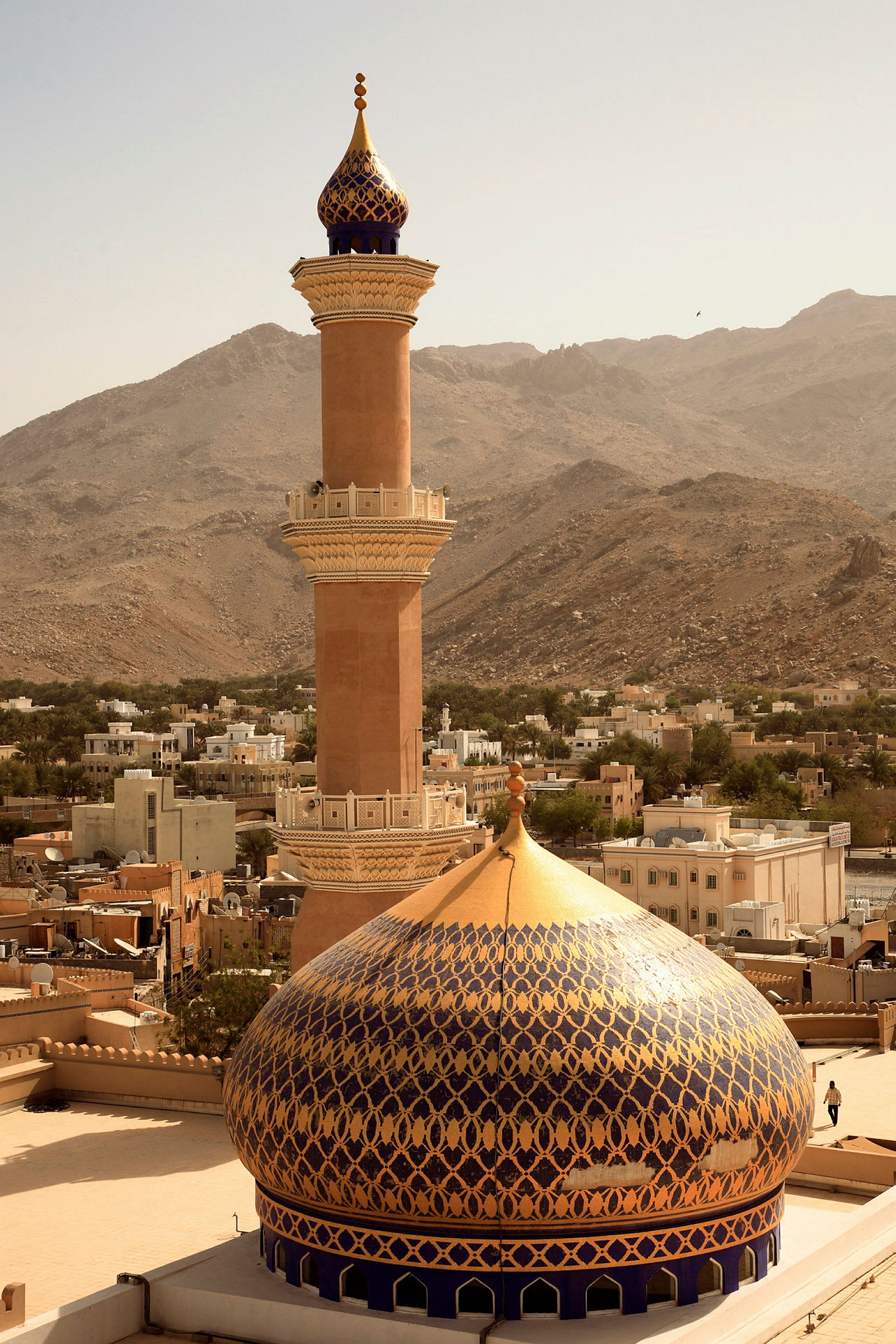 En minaret och en kupol med dekorativa mönster, omgivna av byggnader och berg. (AI-genererad text)