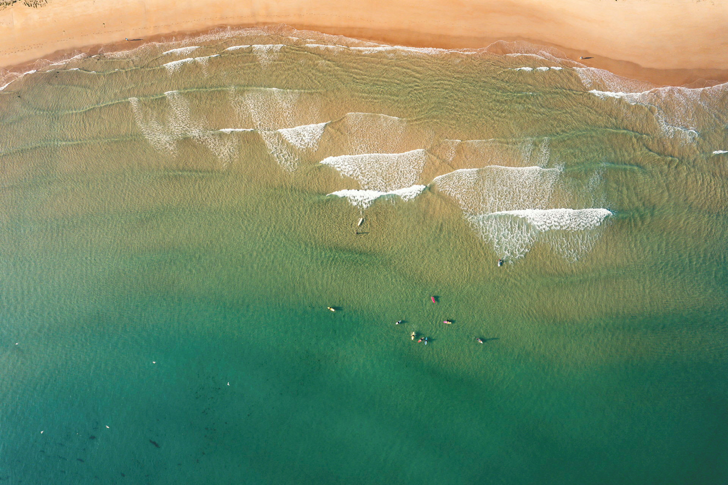 Luftvy av en strand med ljus sand, havsvågor och personer i vattnet. (AI-genererad text)