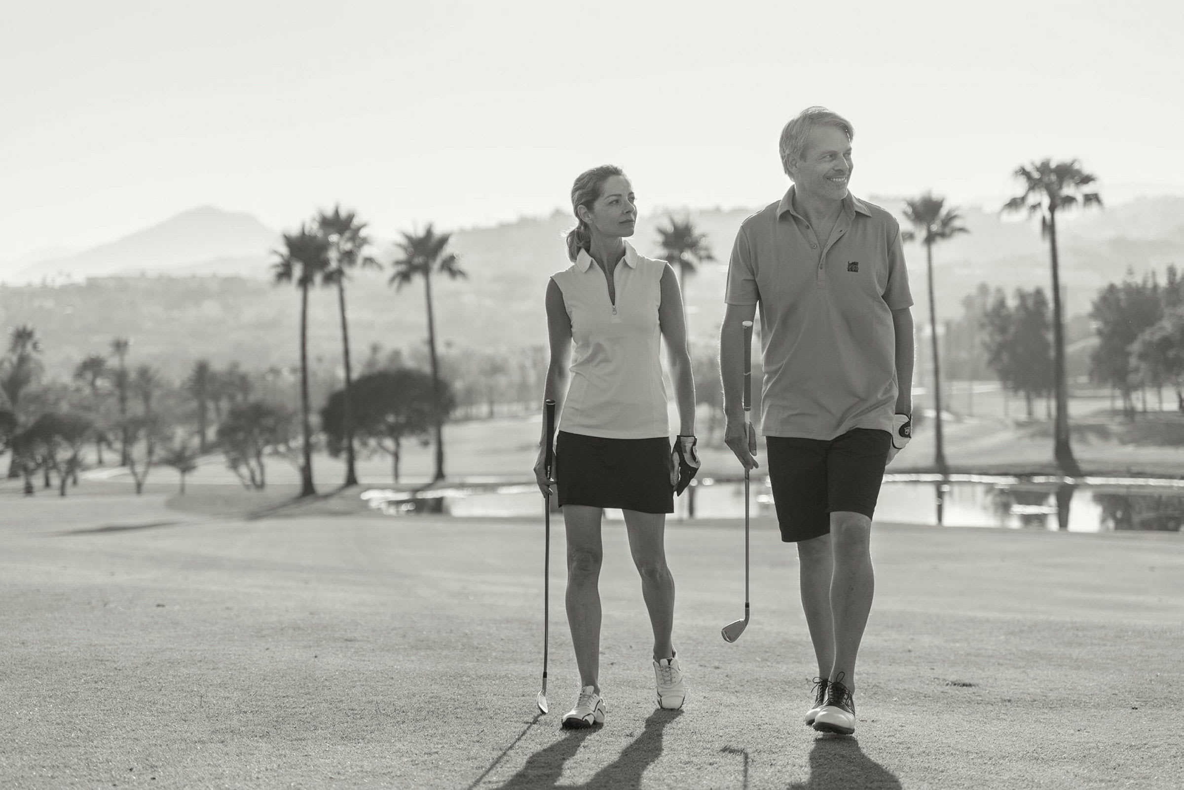 Två personer går med golfklubbor på en golfbana mot en bakgrund av palmer och berg. (AI-genererad text)