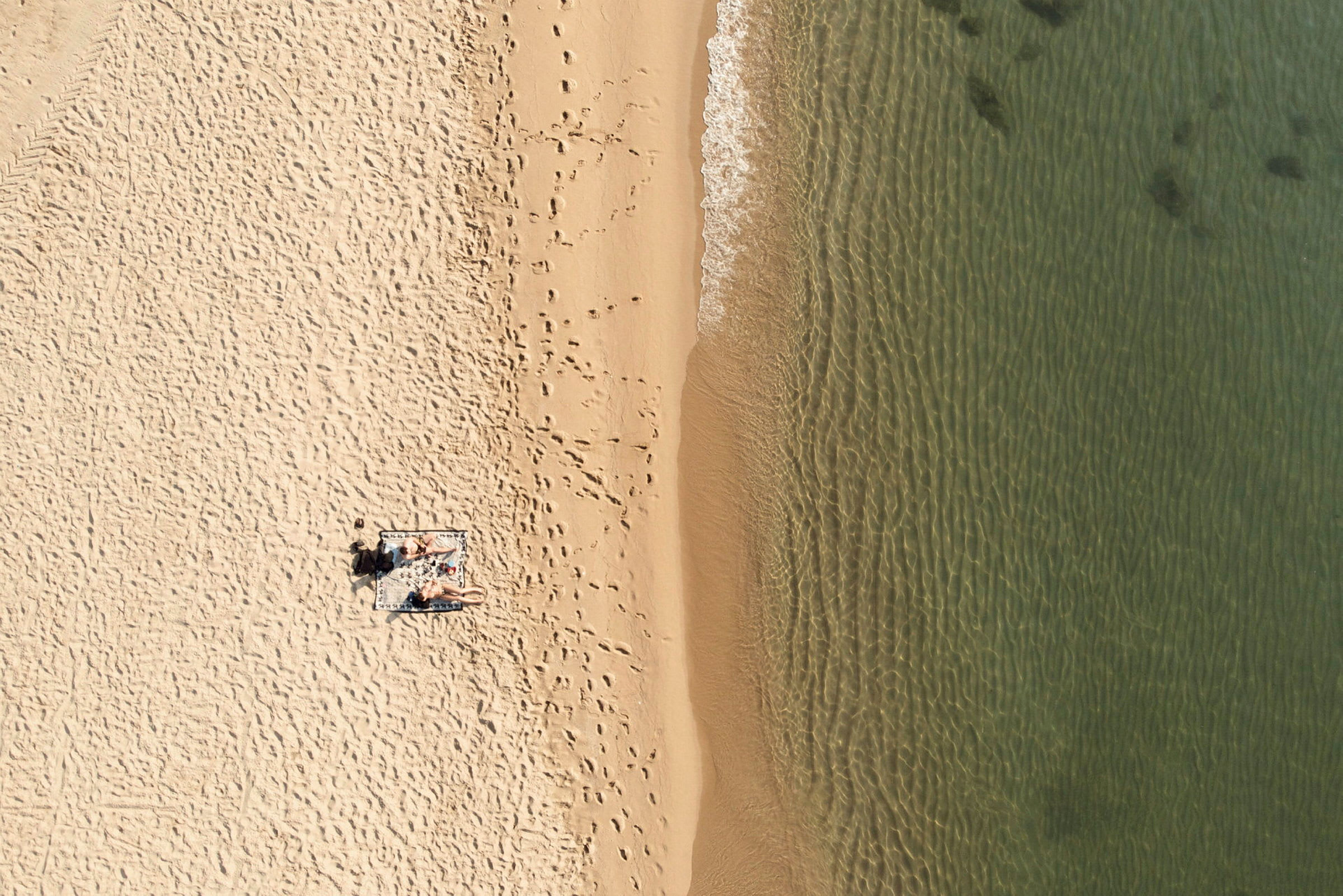 Flygbild av en strand med sand, vatten och personer liggande på en handduk. (AI-genererad text)