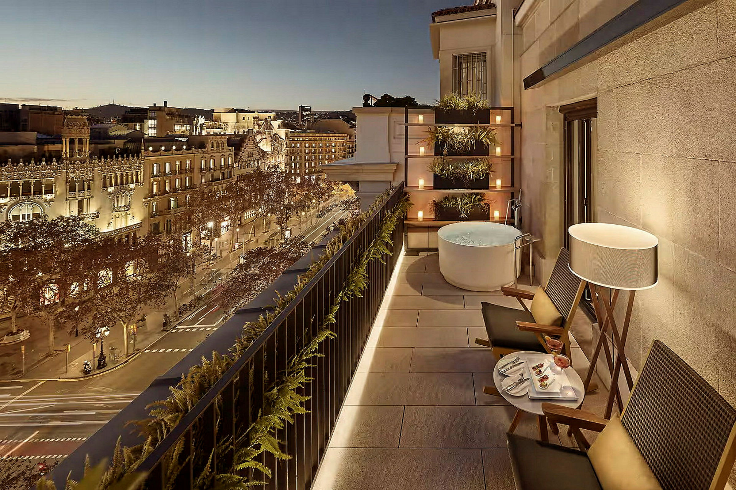 En terrass med ett upplyst bord, stolar och en jacuzzitunna, utsikt över en stad på kvällen. (AI-genererad text)