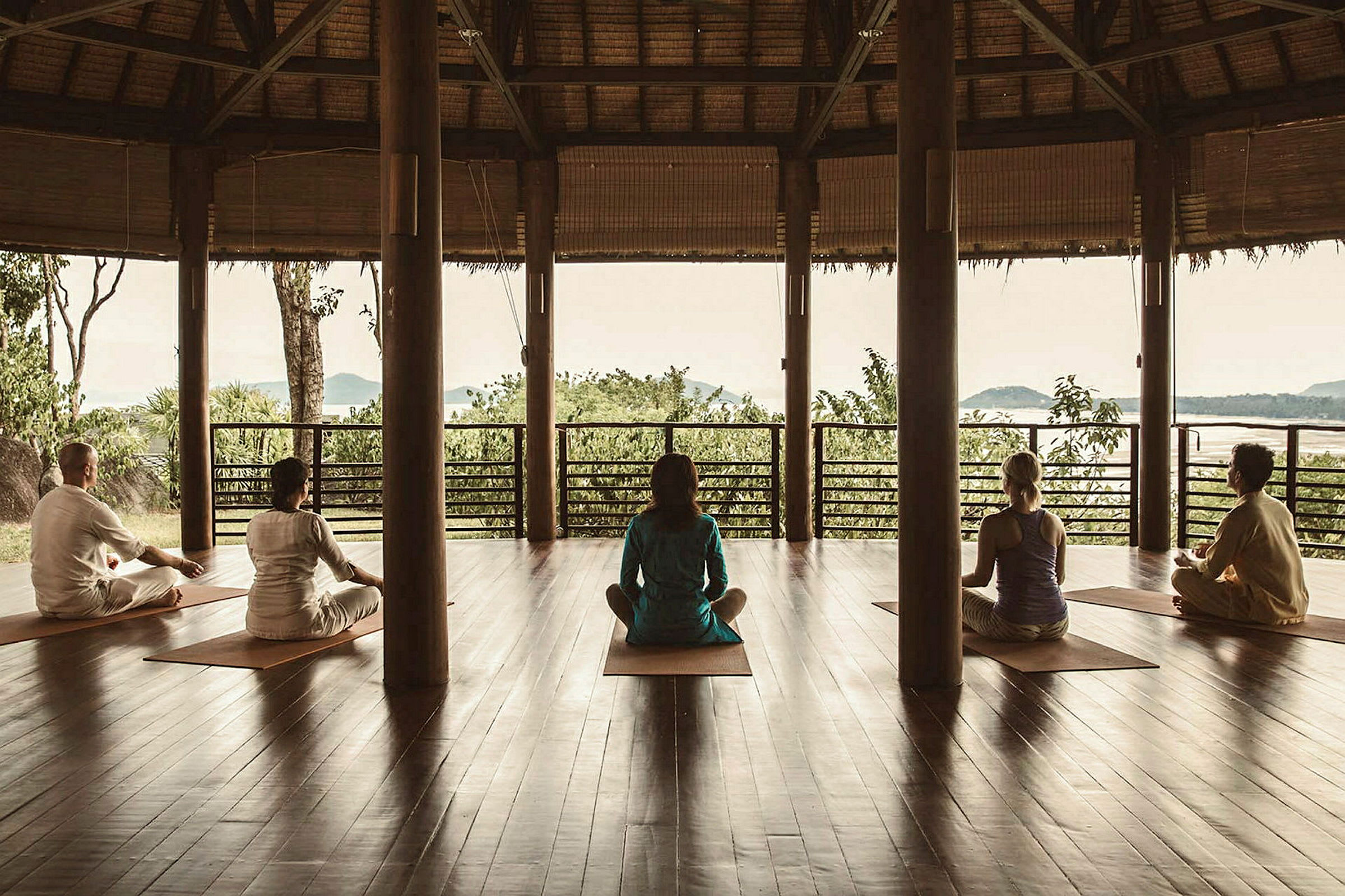 Personer sitter i meditation på yogamattor i en öppen byggnad med trästolpar och utsikt över naturen. (AI-genererad text)