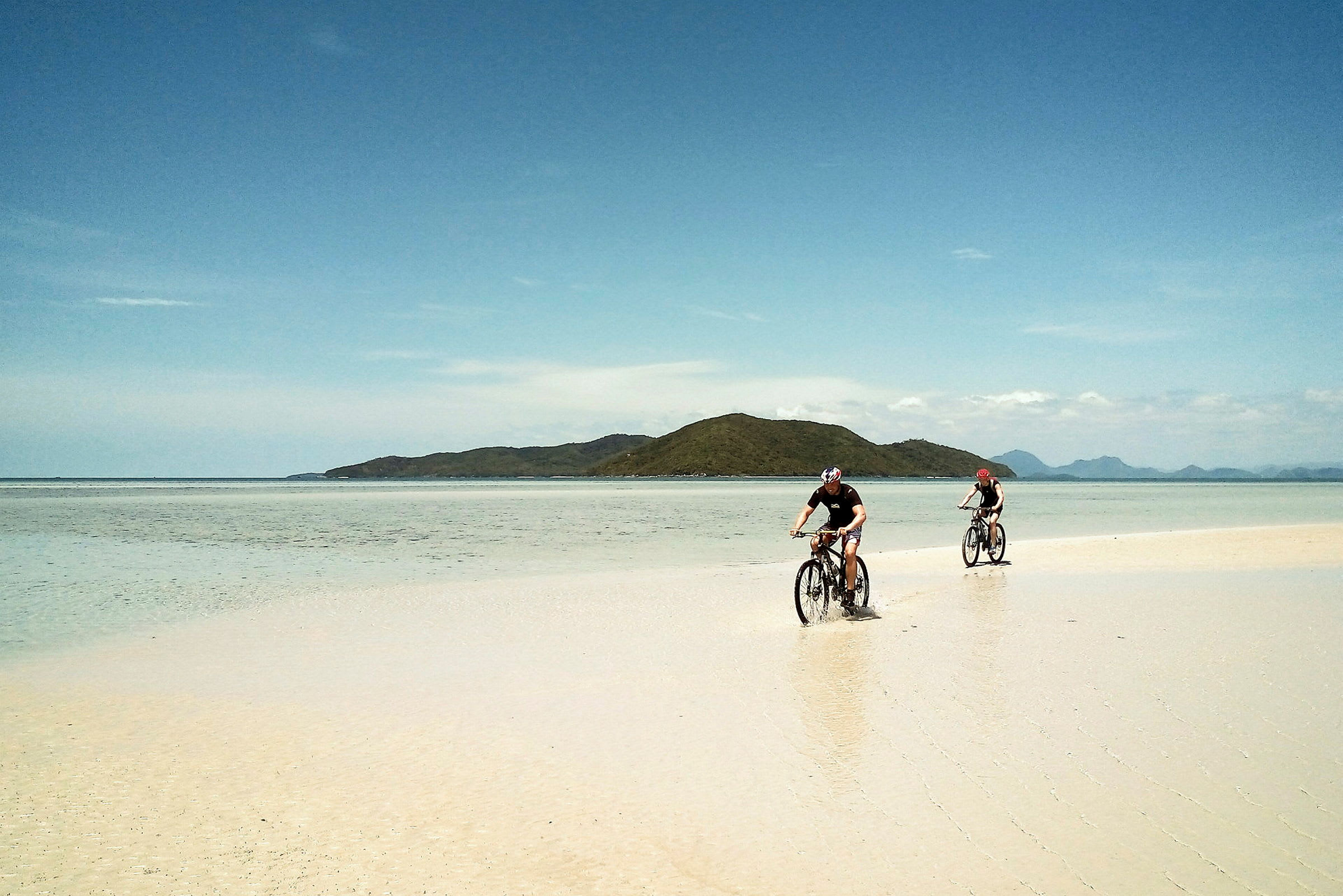 Två personer cyklar på en strand med hav och berg i bakgrunden. (AI-genererad text)