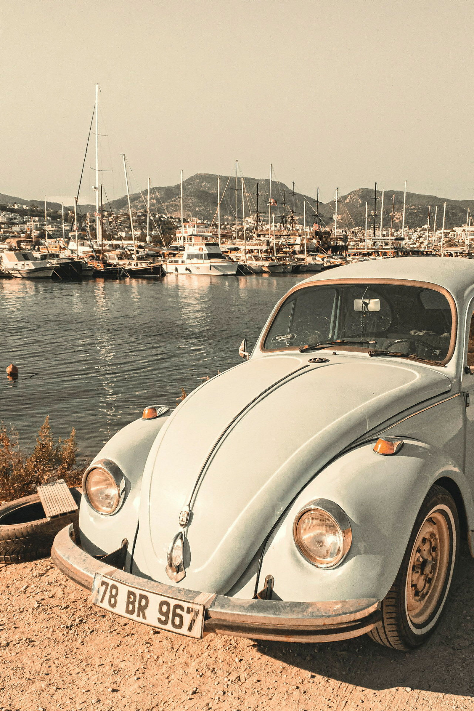 En vintage bil står parkerad vid en småbåtshamn med segelbåtar och berg i bakgrunden. (AI-genererad text)