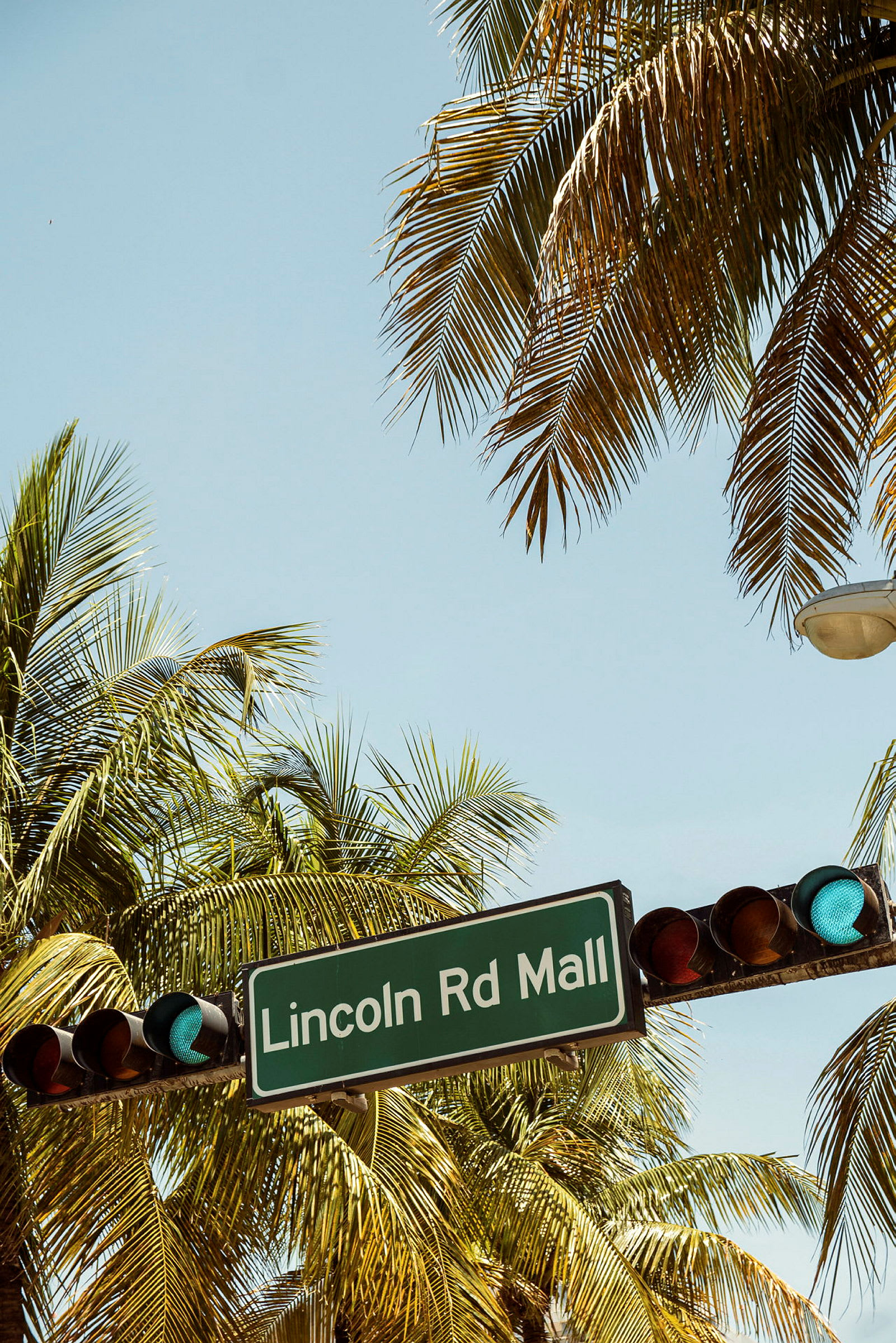 Trafiksignal och skylt med texten 'Lincoln Rd Mall' omgivna av palmer under en klar himmel. (AI-genererad text)
