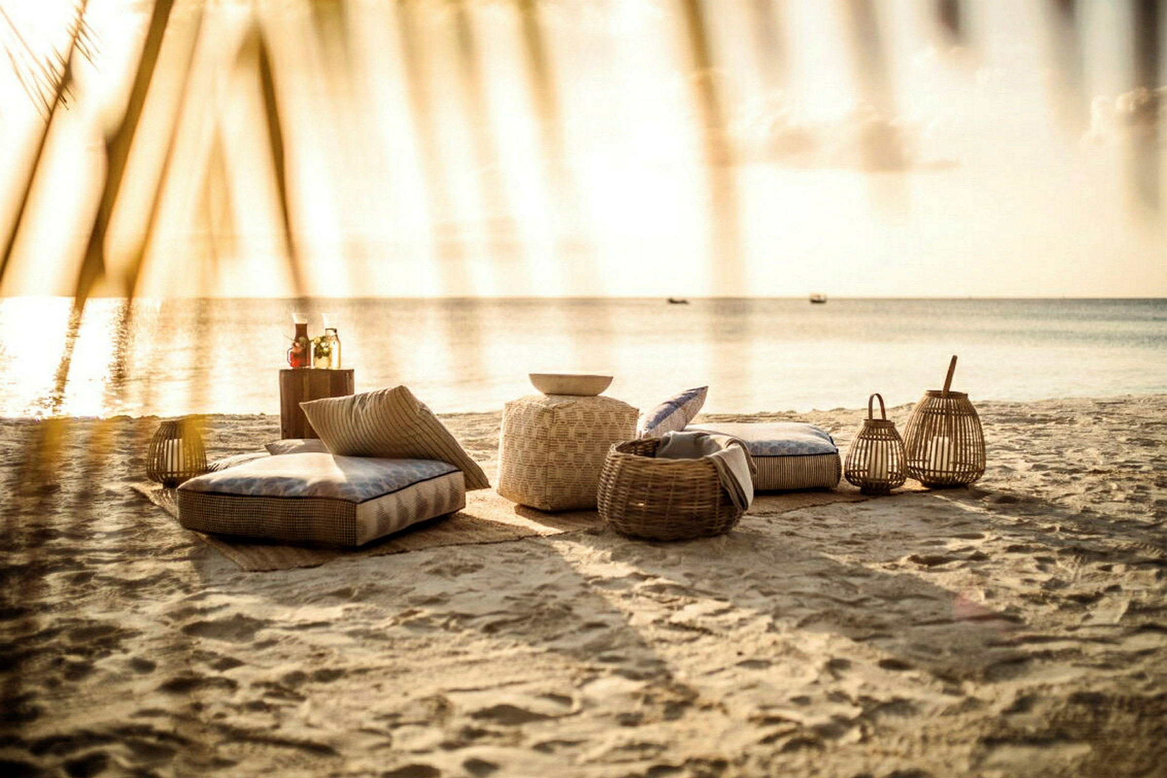 Strand med kuddar, korgar och lyktor vid solnedgång. (AI-genererad text)